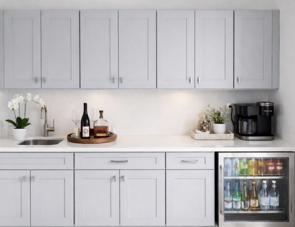 Modern kitchen with white cabinets, black coffee maker, bottles of wine and alcohol, glass of wine, potted orchid, and mini fridge with drinks.