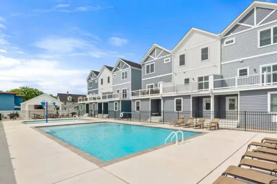 Residential apartment complex with a swimming pool, lounge chairs, and multi-story buildings with balconies under a blue sky.