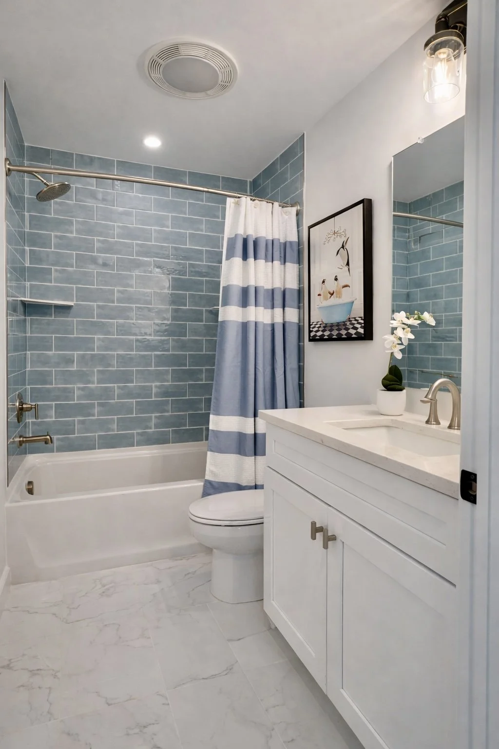 Bathroom with blue tiled shower, white vanity with a potted orchid, framed artwork, and a toilet, featuring a blue and white striped shower curtain.