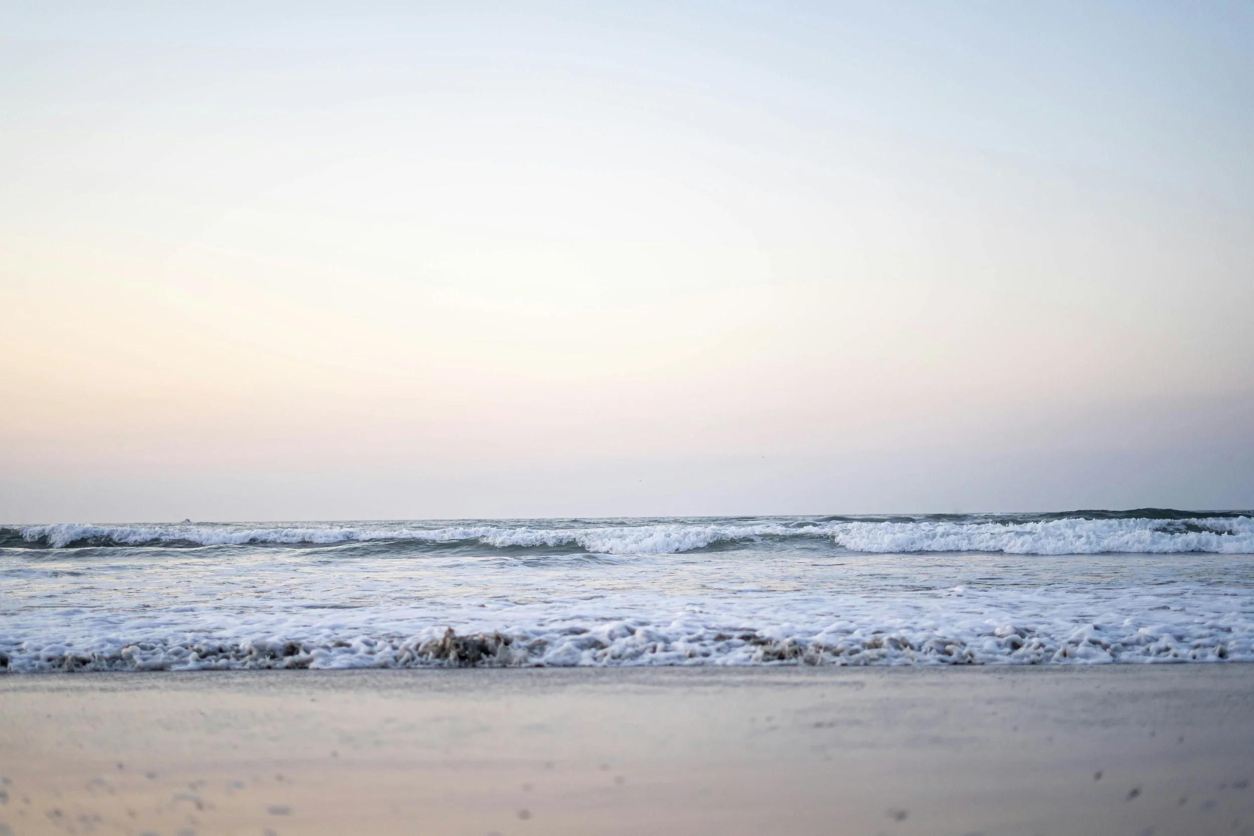A scenic view of the ocean with gentle waves rolling onto the beach at sunset or sunrise, with a cloudy sky.