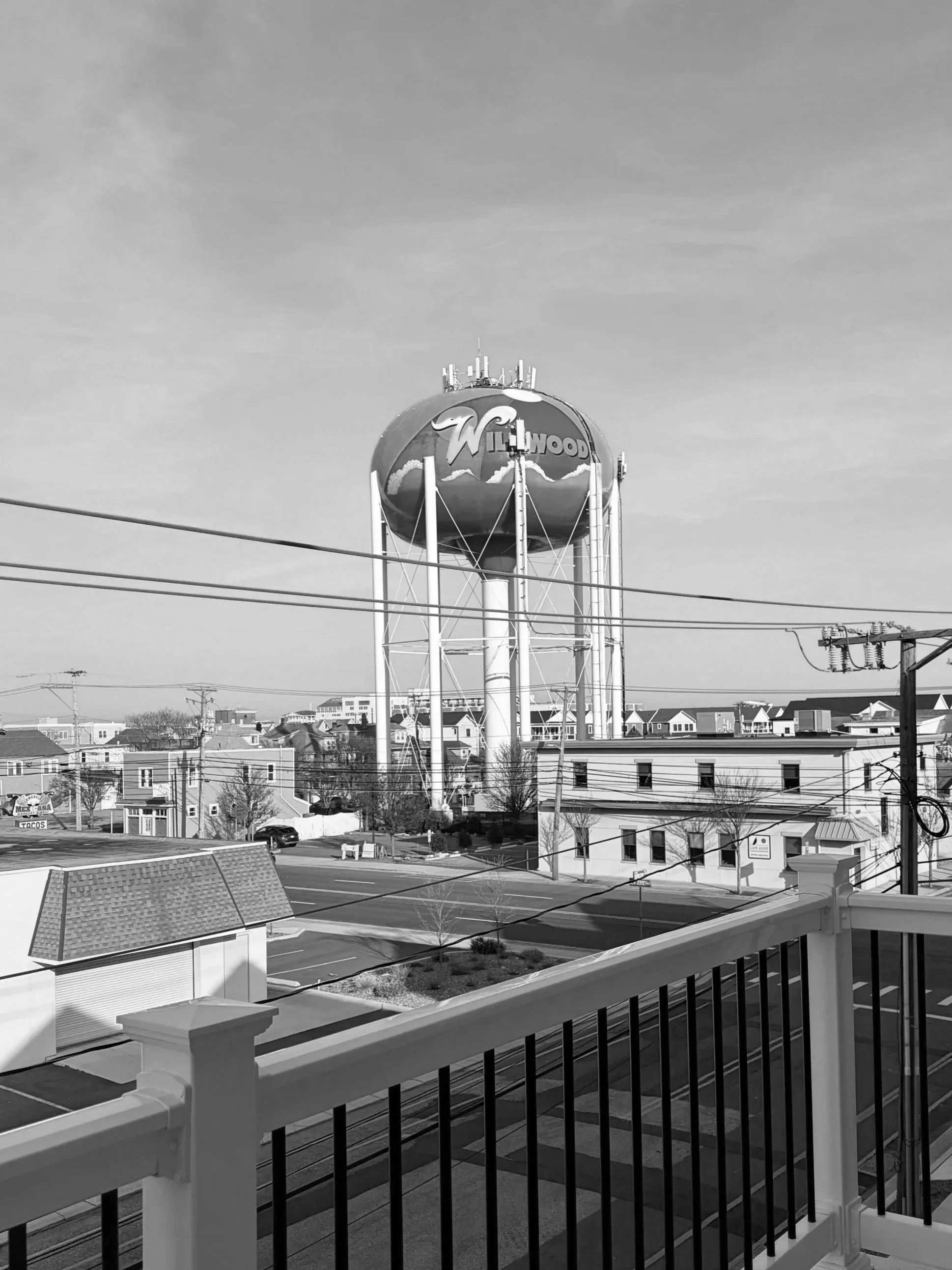 Black and white photo of a water tower with the words 'Willowood' and a mountain and wave design, surrounding a cityscape with power lines, buildings, and rooftops, viewed from a balcony with a railing.