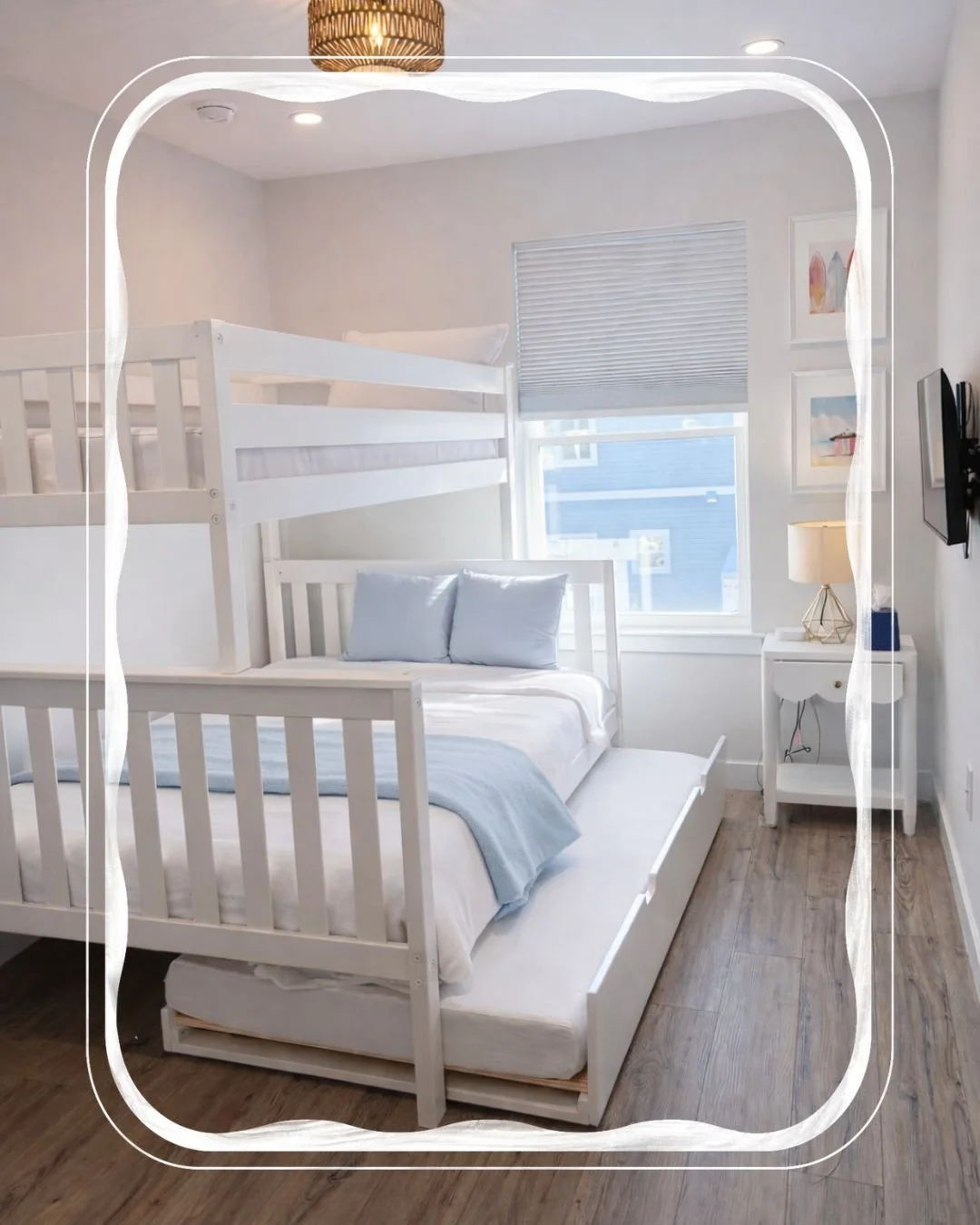 A bedroom with a white bunk bed, a white nightstand with a lamp, and a small bed underneath. The room has a window with blinds, several framed pictures, and wooden flooring.