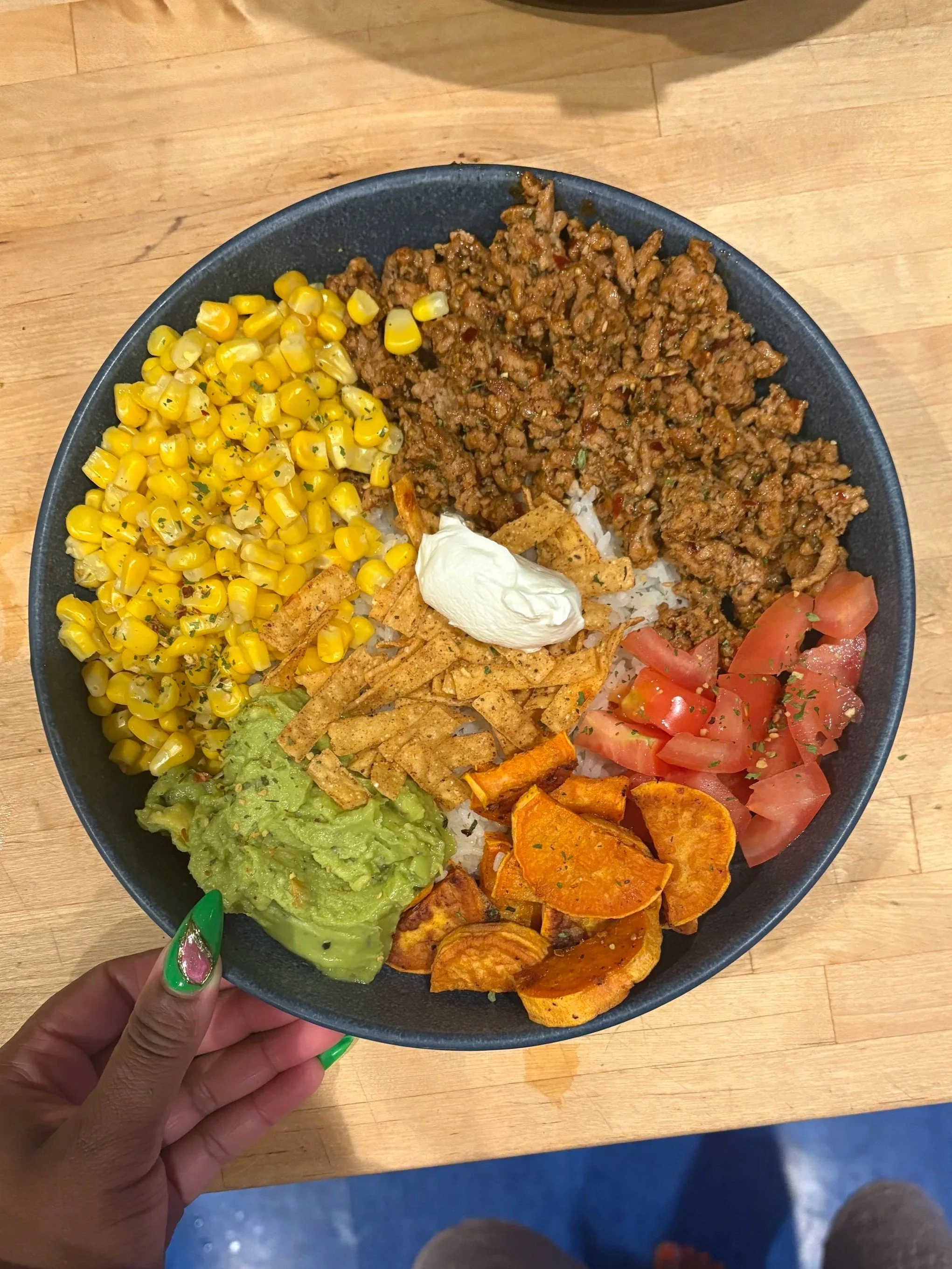 A hand holding a black bowl filled with corn, seasoned ground meat, diced tomatoes, guacamole, cooked sweet potatoes, shredded chicken, and rice with toppings.