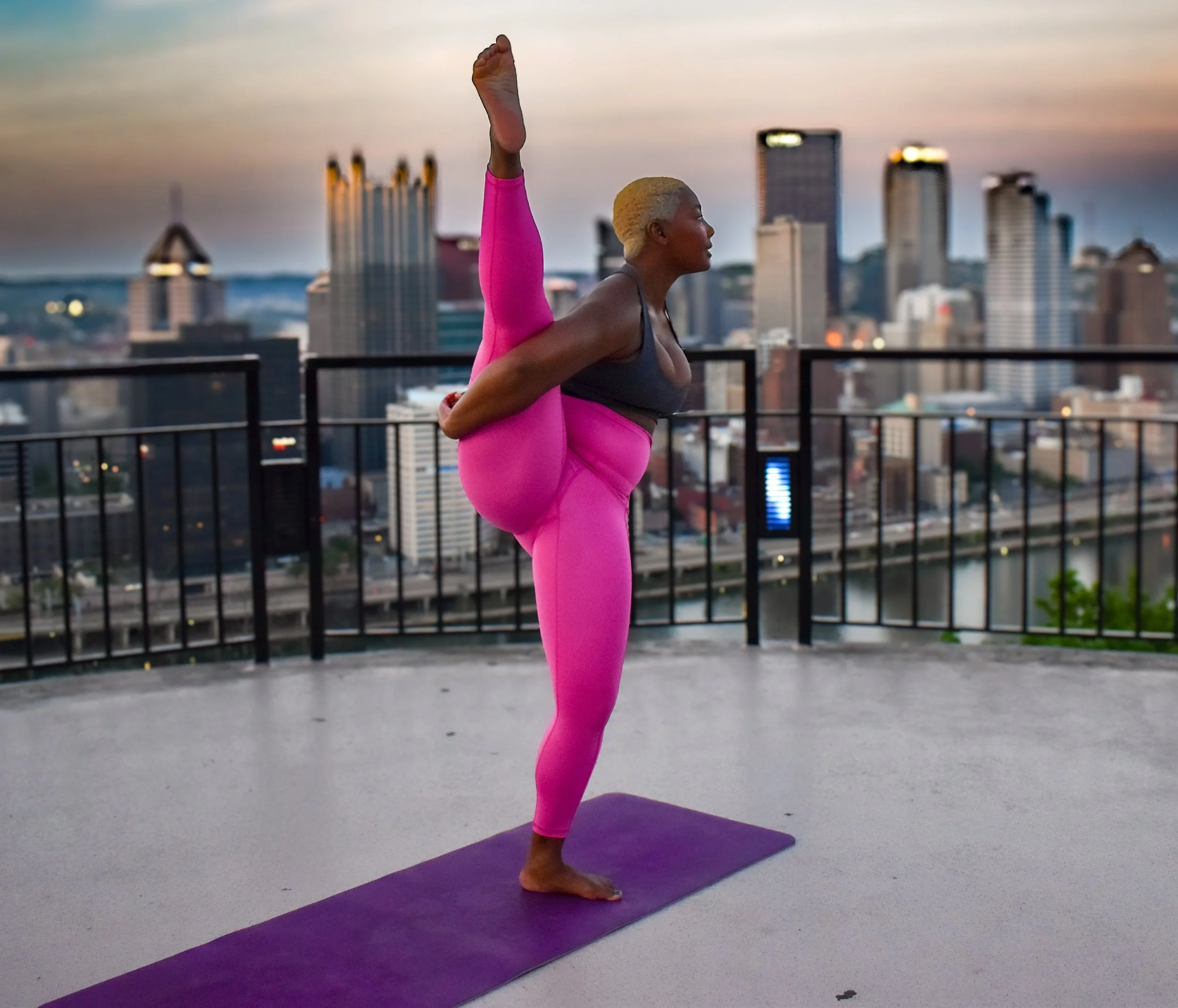 A woman with short blonde hair practicing yoga on a purple mat on a rooftop at sunset, performing a standing split pose with one leg raised straight up and her foot resting near her head, overlooking a city skyline.