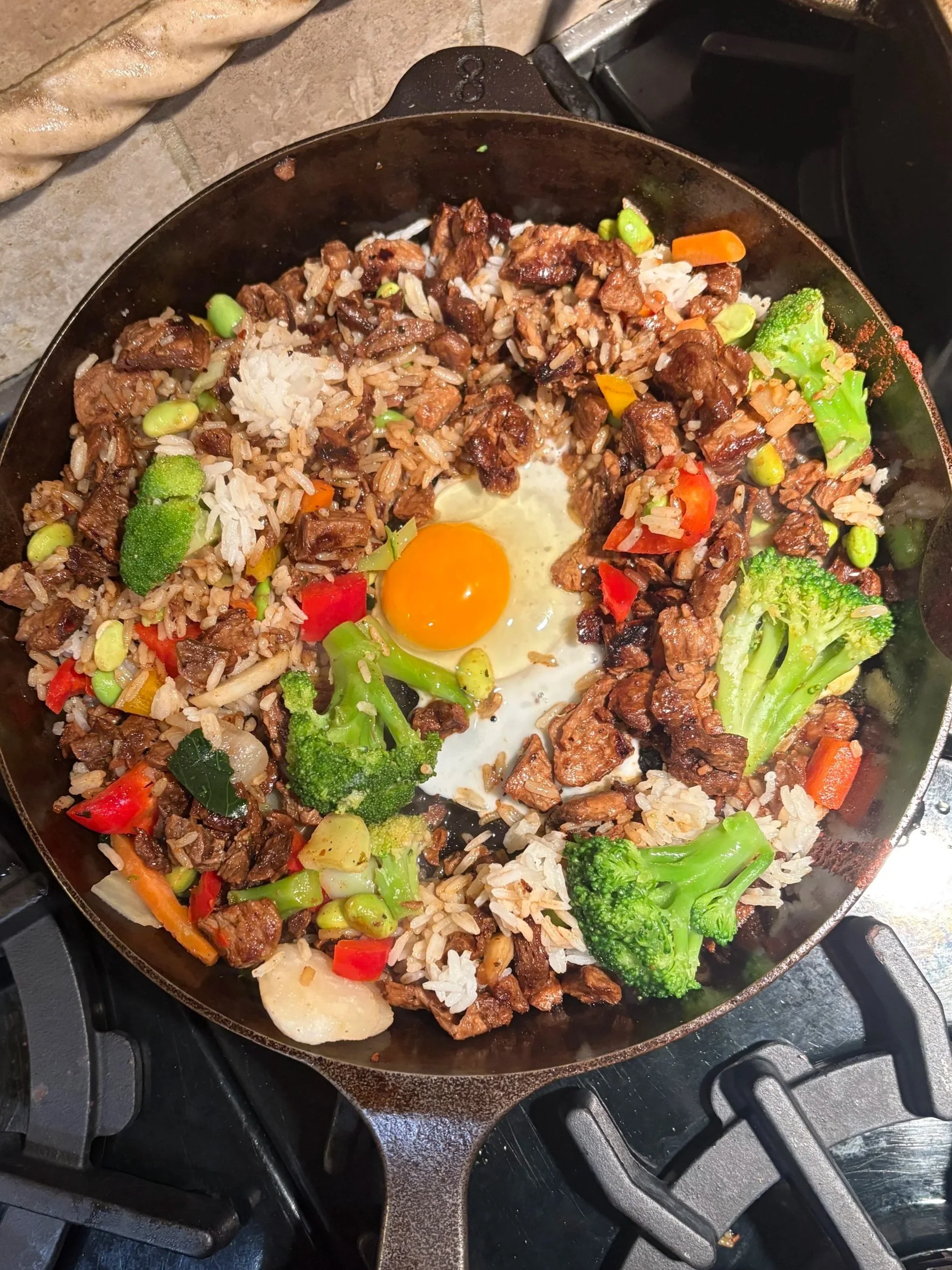 A skillet on a stovetop containing cooked rice, beef chunks, sautéed vegetables including broccoli, red and yellow bell peppers, carrots, and an egg with a runny yolk.