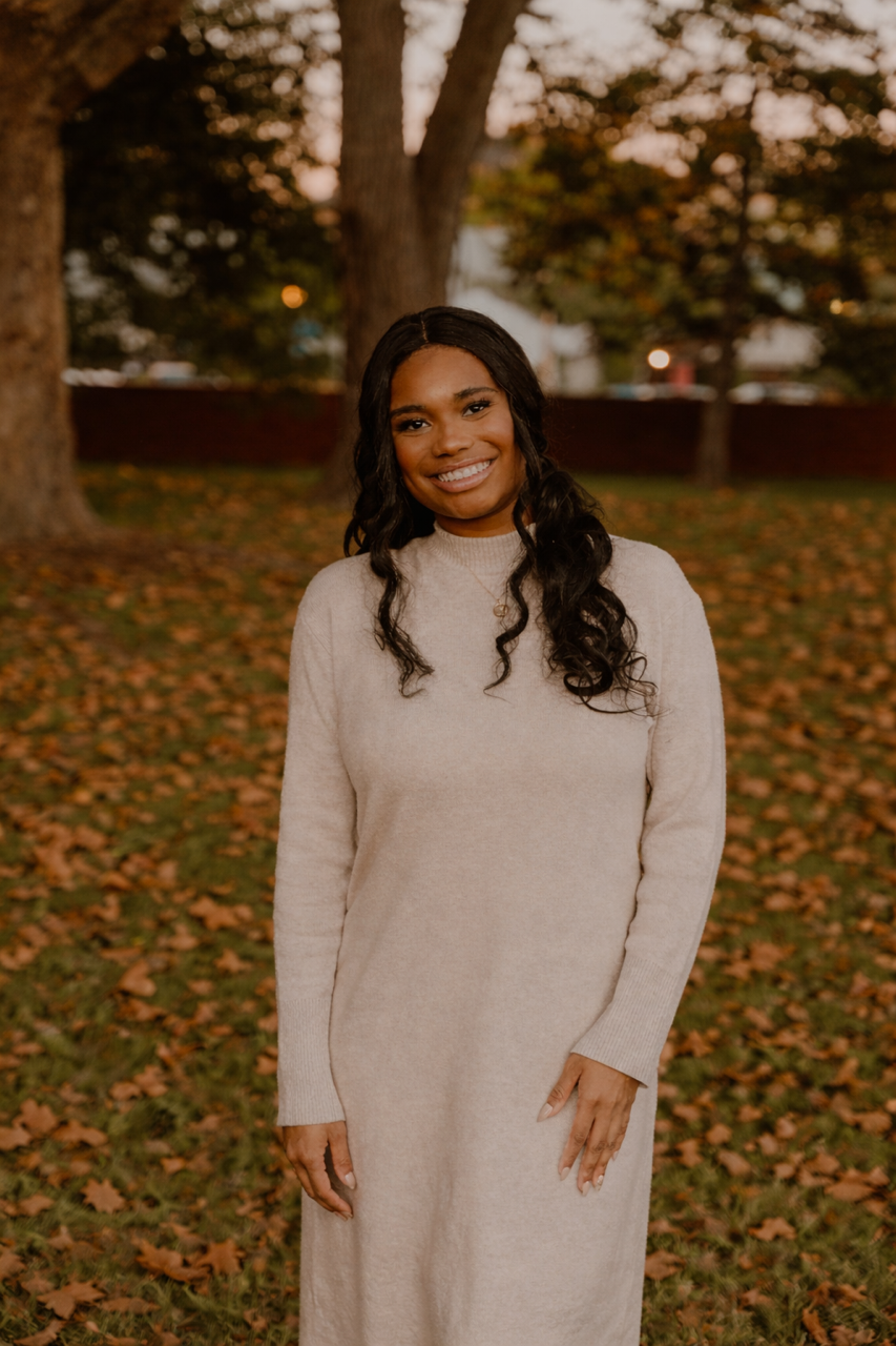 A woman in a beige sweater dress standing outdoors during fall, with fallen leaves on the ground and trees in the background.