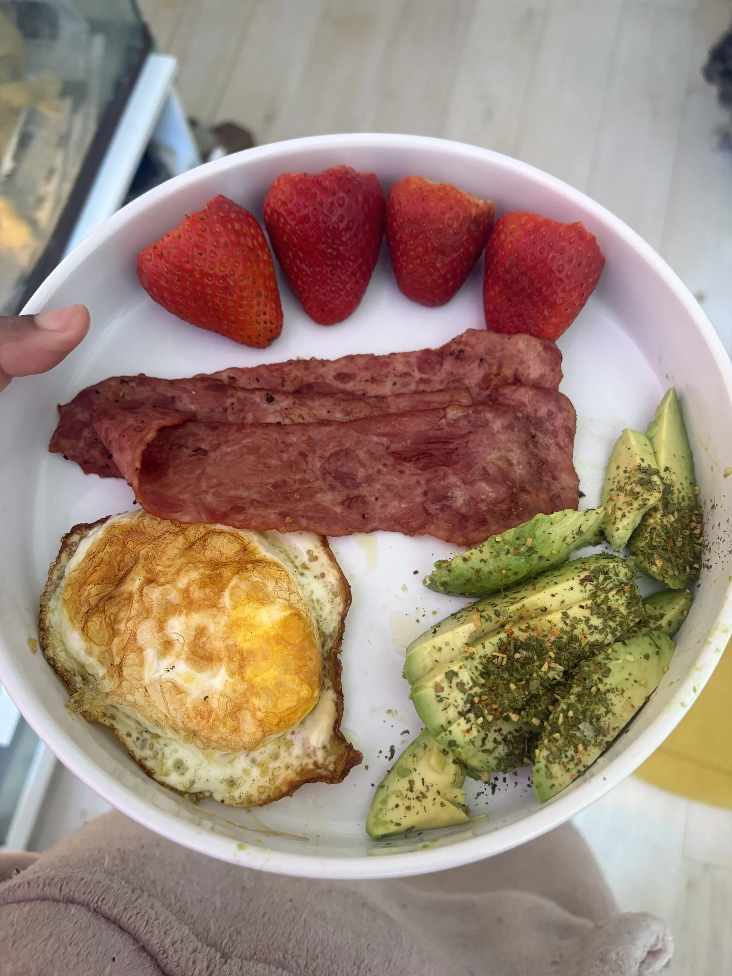 Bowl of breakfast food with strawberries, fried egg, slices of avocado, and cooked meat.