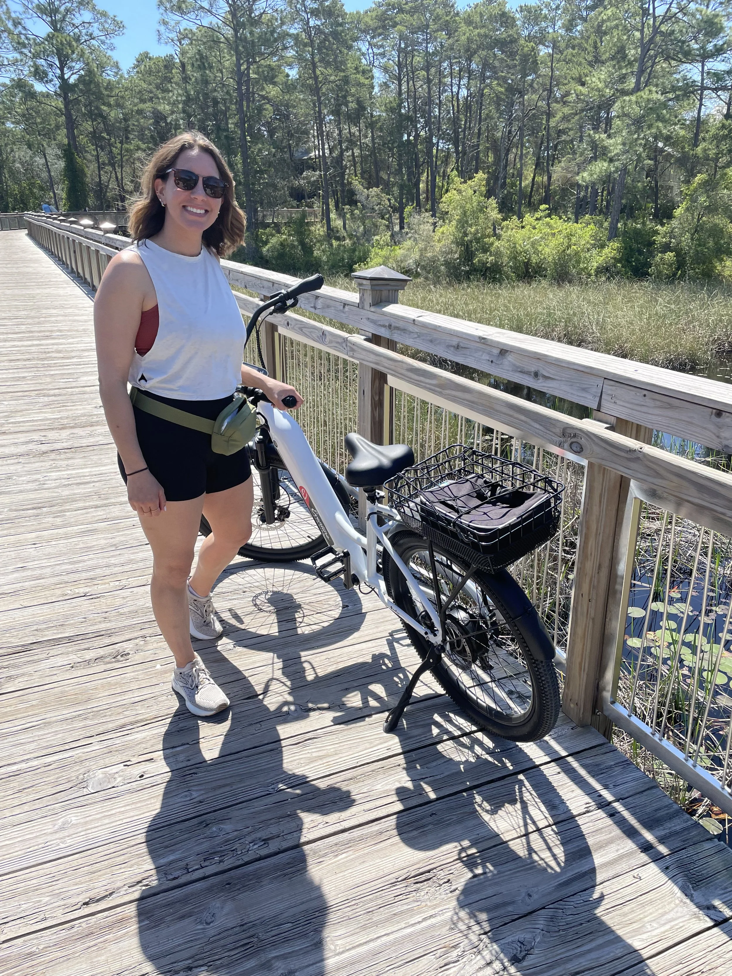 A woman with sunglasses, wearing a white sleeveless shirt and black shorts, standing on a wooden bridge next to an electric bicycle with a basket, in a natural area with trees and water in the background on a sunny day.