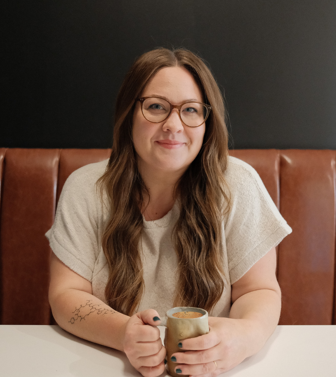 A woman with long wavy hair, glasses, and a white shirt, sitting at a table in a cafe, holding a mug of coffee, with a dark wall and brown leather booth in the background.