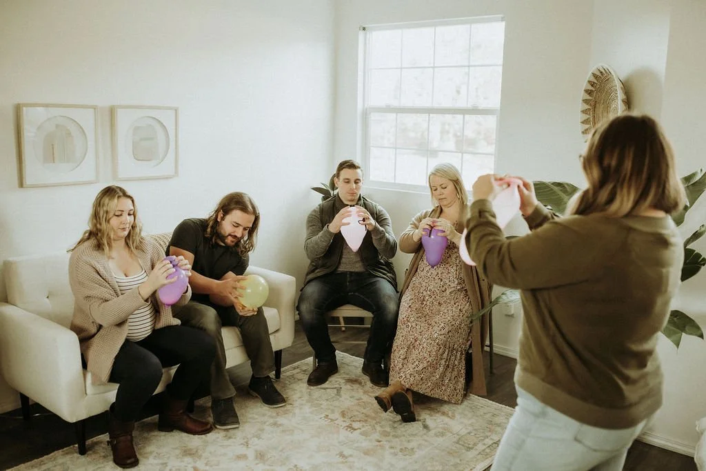 Five people sitting and standing in a bright living room, blowing up pink, yellow, and purple balloons.