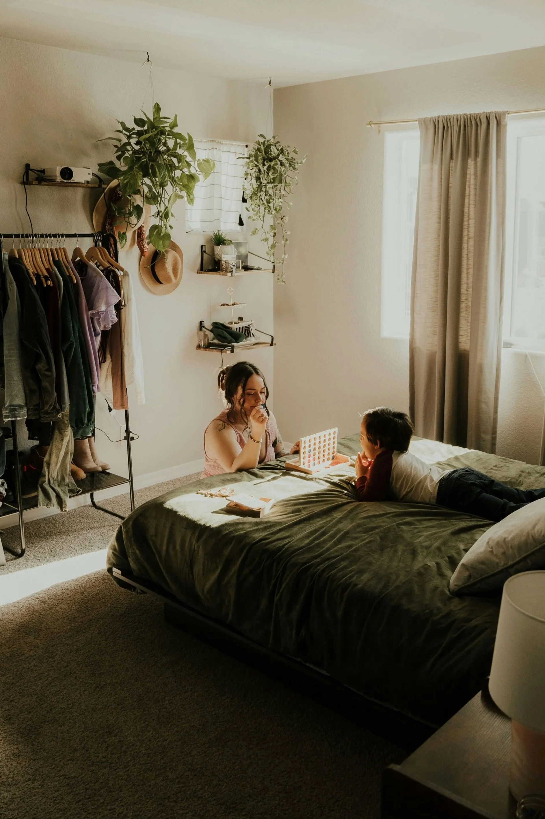 A bedroom with a large bed, one women playing a game with a child, a clothing rack, hanging plants, and a window with curtains.