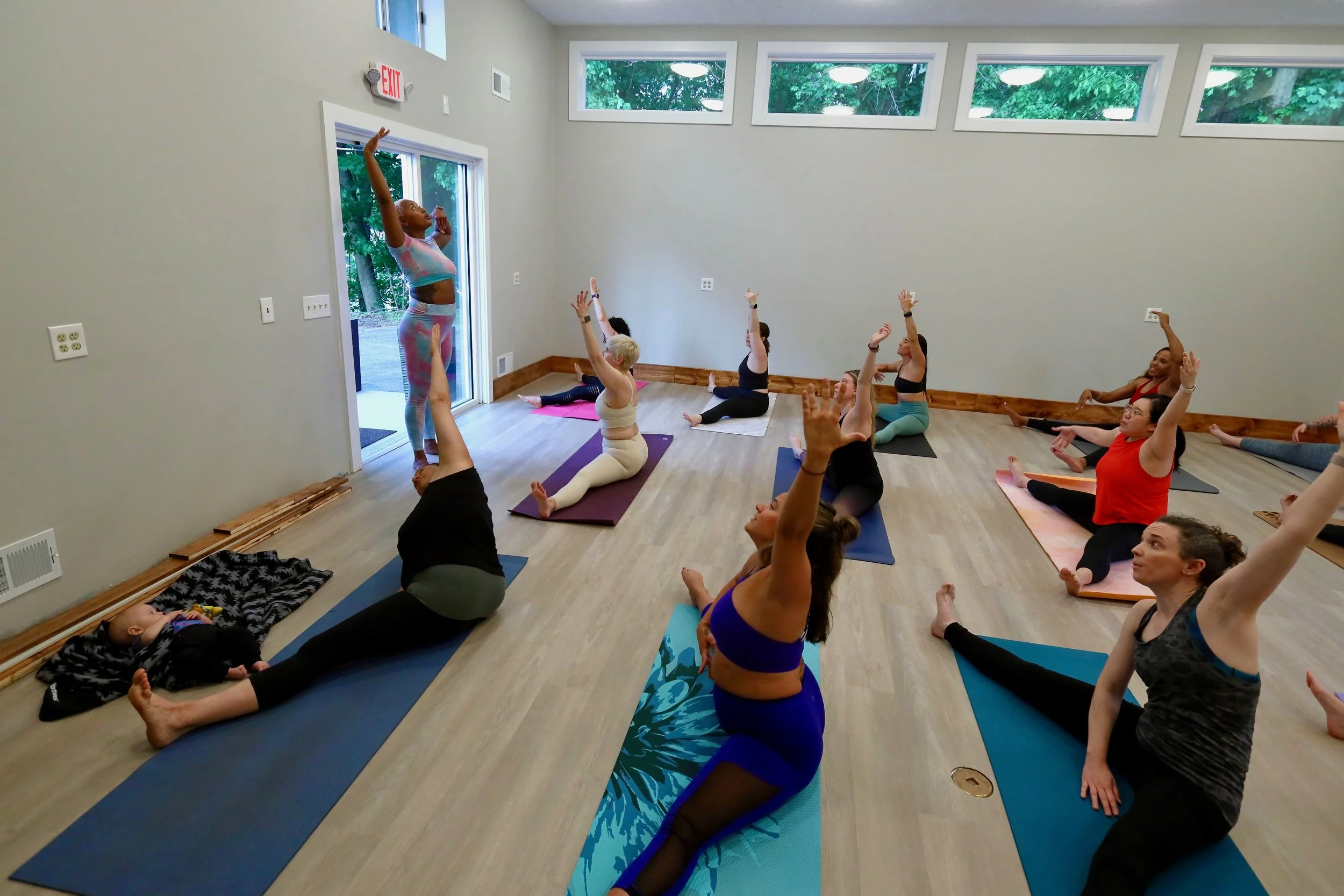 A group of people participating in a yoga class in a spacious, well-lit room with wooden floors and large windows, led by an instructor demonstrating a pose near the door.