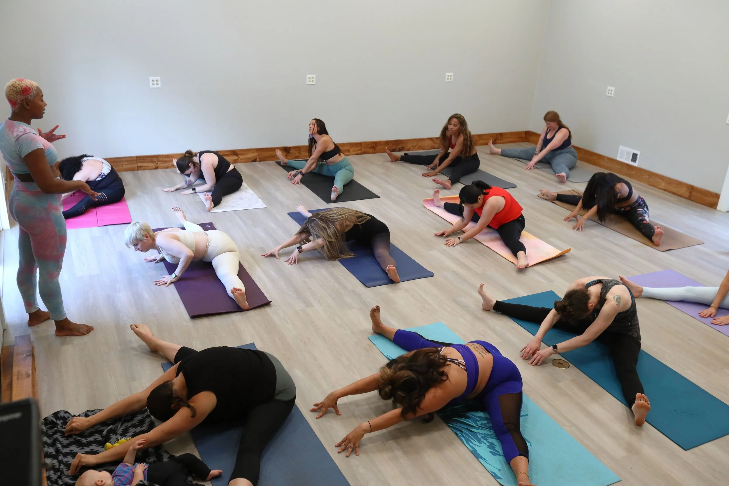 A group of women participating in a Prenatal yoga class, performing stretches on their mats in a spacious room with light-colored walls.
