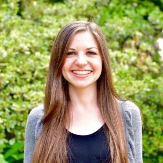 Young woman with long brown hair smiling outdoors in front of green foliage.