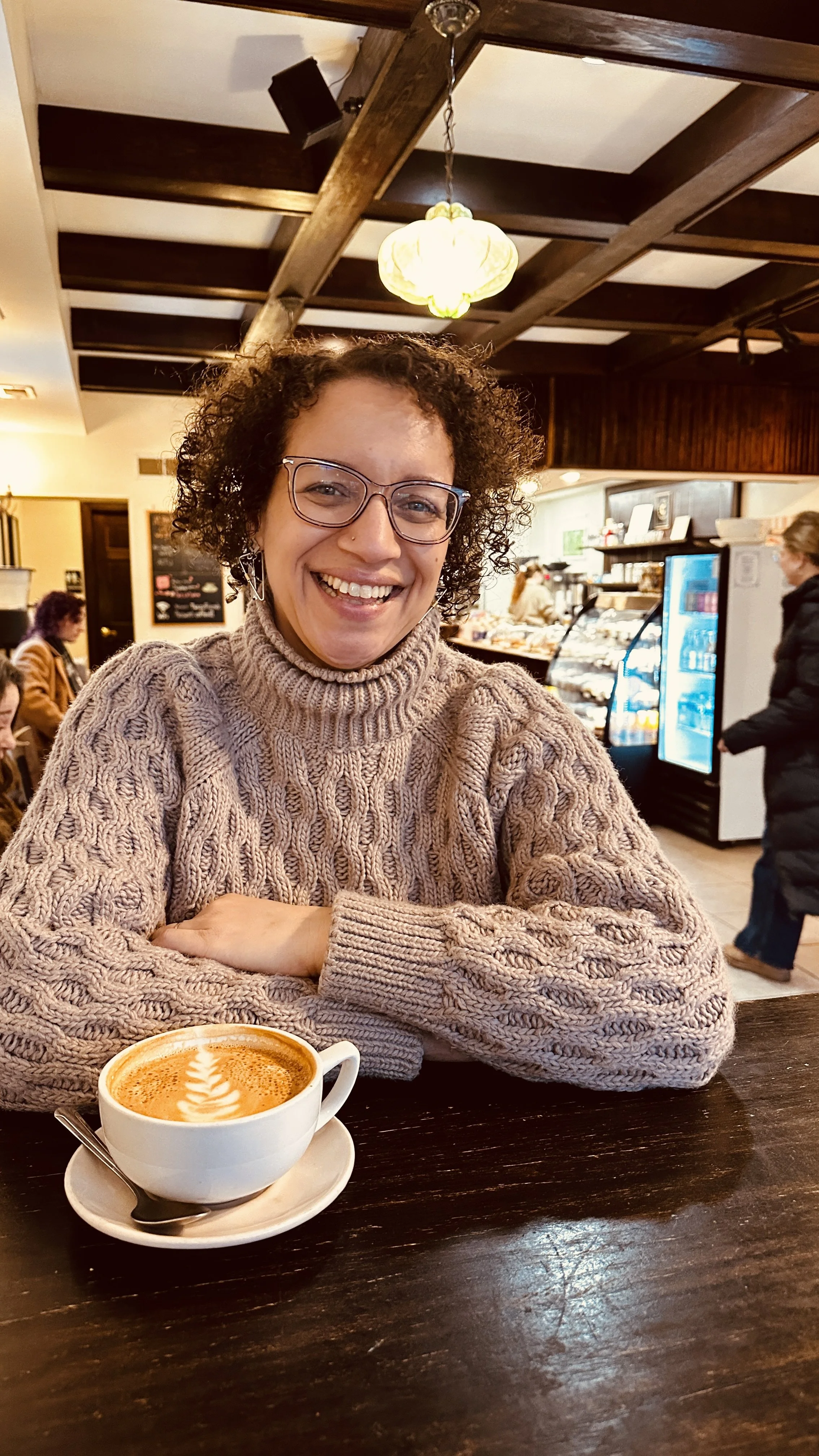 A woman with curly hair, glasses, and a beige cable-knit sweater sitting at a table with a cup of coffee that has latte art. She is smiling inside a cozy coffee shop with other customers and a counter with baked goods in the background.