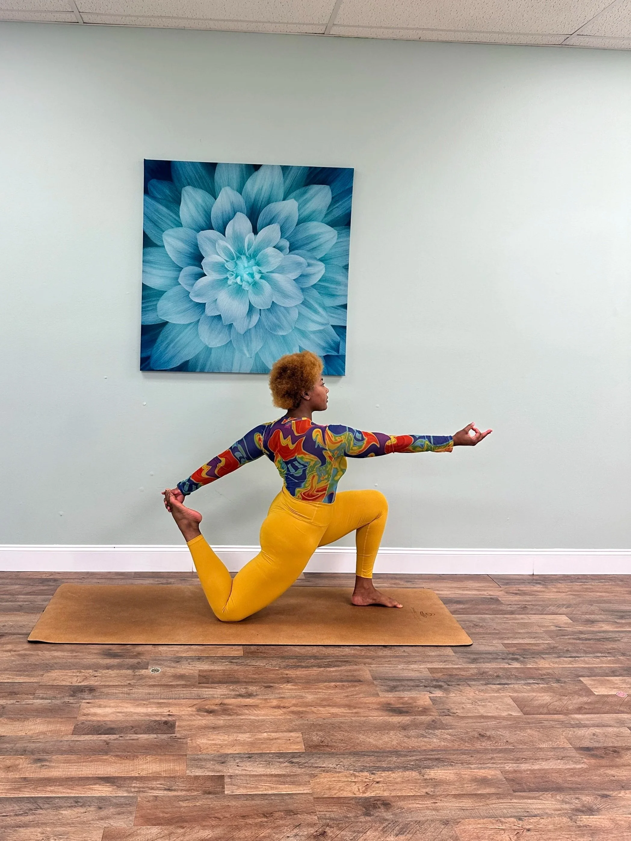 A Black woman practicing yoga in a lunge pose on a brown mat in a room with light-colored walls and a large blue flower painting on the wall.