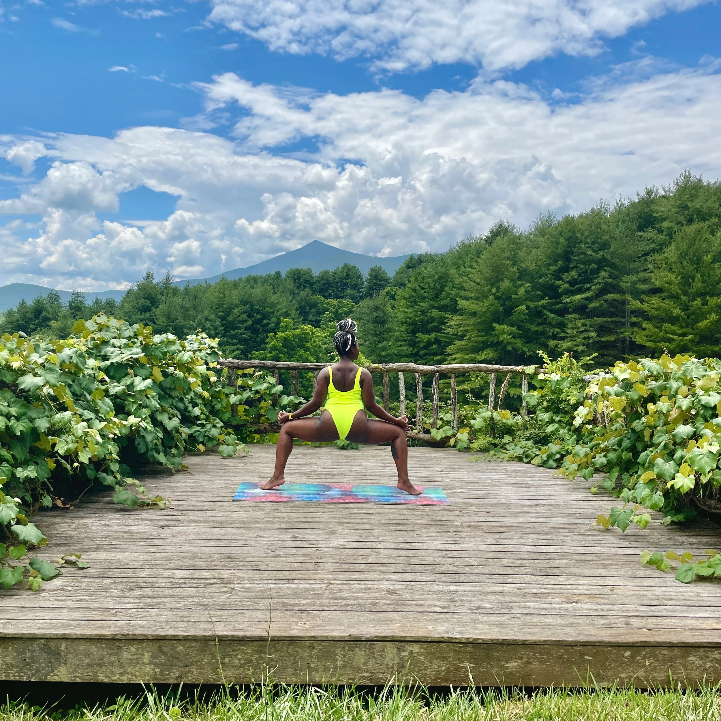 A Black woman in a yellow outfit practicing yoga on a wooden deck outdoors, surrounded by green plants and trees, with mountains and a blue sky with clouds in the background.