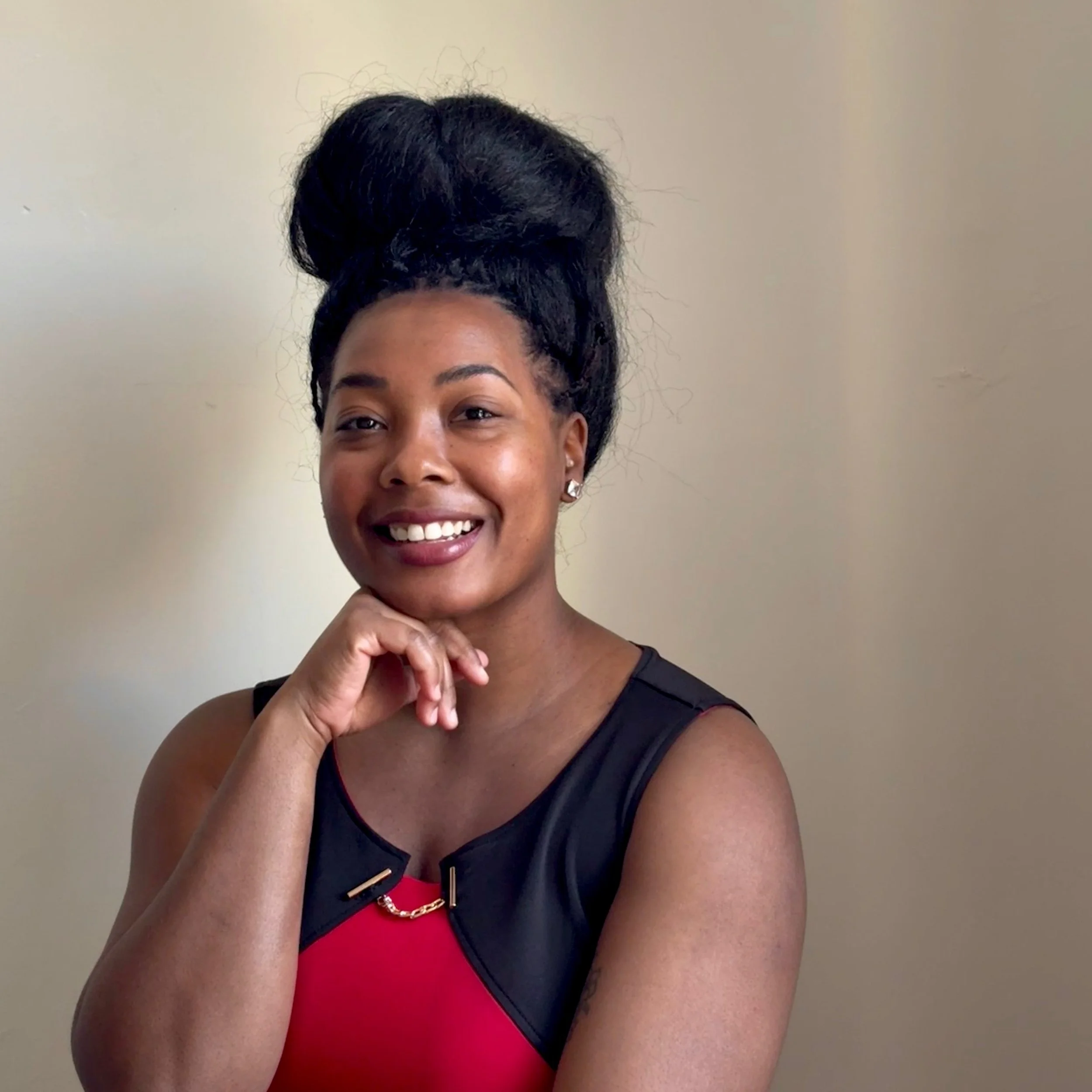 Close-up of a smiling Black woman with dark hair styled in an updo, wearing a black and red sleeveless top and a gold chain necklace, against a plain beige background.