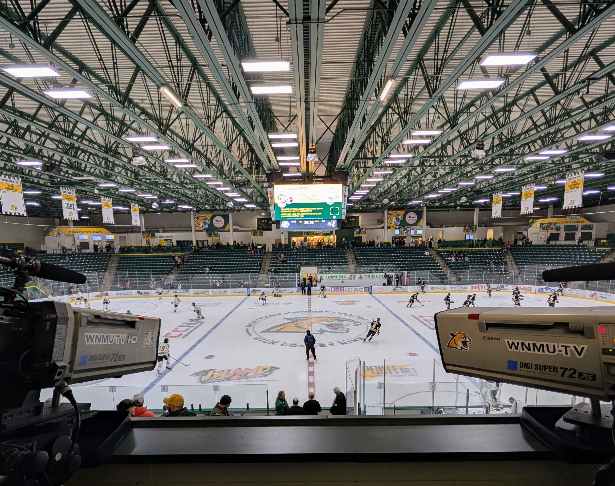 An indoor ice hockey rink with players warming up or practicing, surrounded by seating for spectators and media cameras in the foreground.
