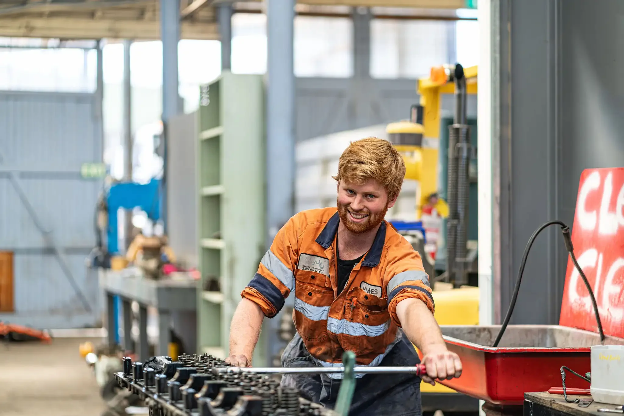 A smiling man with red hair, beard, and blue eyes working in an industrial or workshop setting. He is wearing a high-visibility work jacket with orange and blue colors and is leaning over a workbench, handling tools or machinery, with various equipment and shelves in the background.