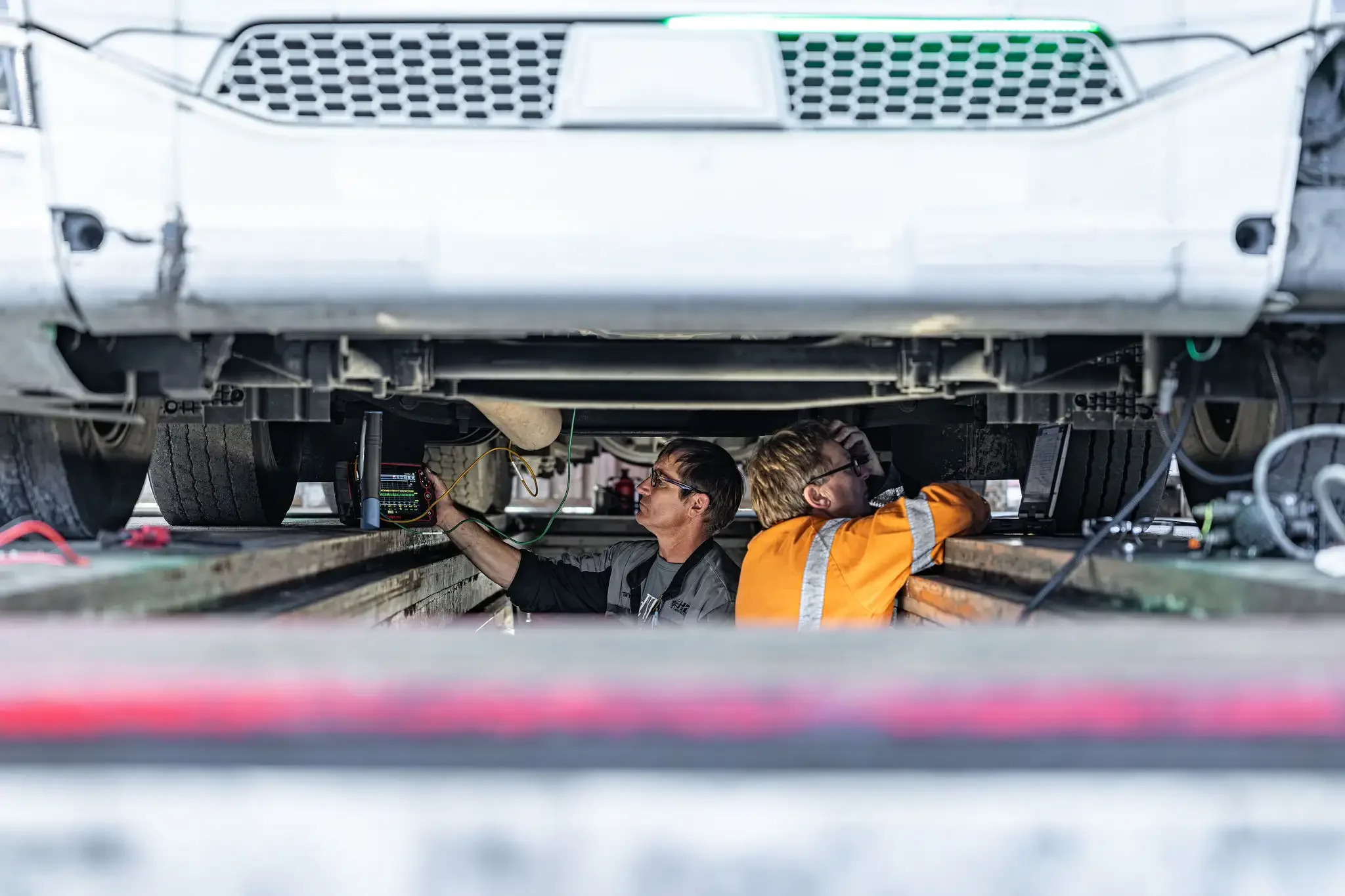 Two vehicle technicians working underneath a large white vehicle, possibly an electric truck, using various diagnostic tools and wires for maintenance or repair.