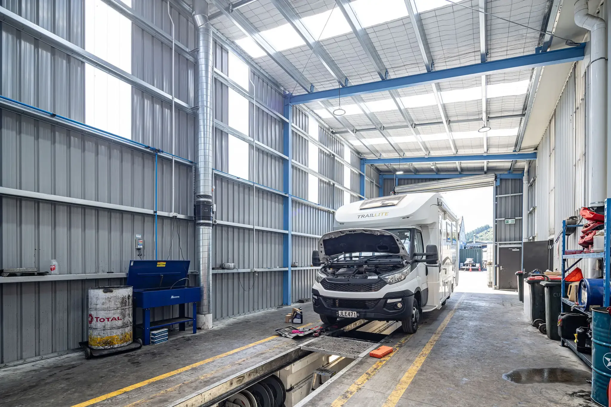 Inside an automotive workshop with a white camper trailer labeled 'TrailLite' and its hood open. The workshop has high ceilings, metal walls, and a car lift with a yellow safety line on the floor. There are various tools and equipment around, including a blue toolbox, an oil drum, and other containers.