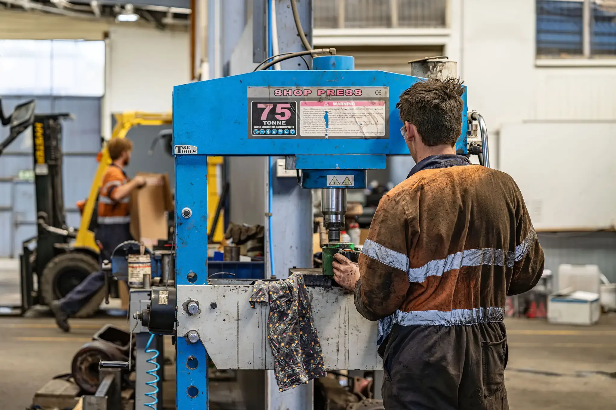 A worker in a brown and orange safety jumpsuit operates a blue industrial machine in a factory setting. In the background, another worker is seen moving a box near a forklift, with various machinery and large windows visible.