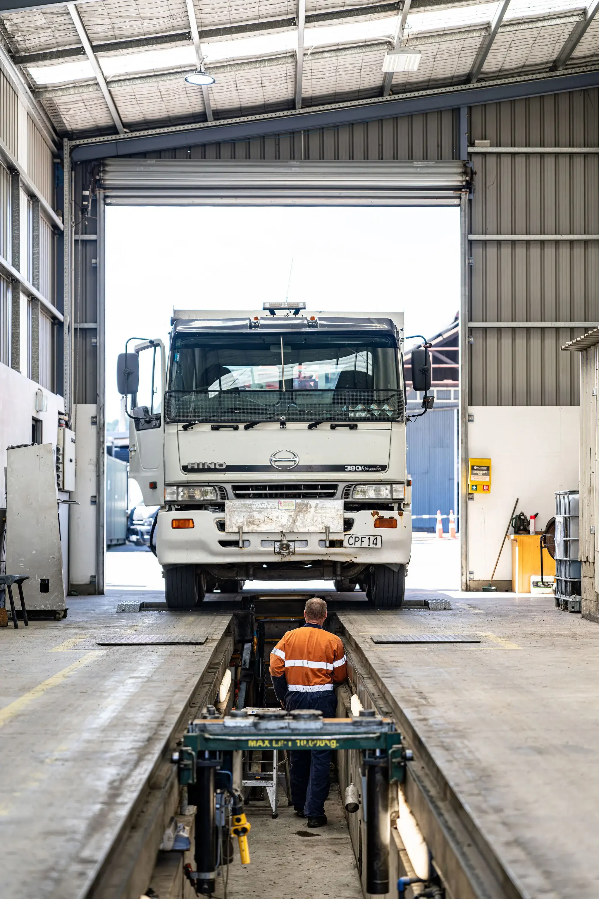 A large white truck inside an industrial garage with a worker inspecting underneath. The garage has a high arched ceiling with metal walls and an open partially rolled-up door.