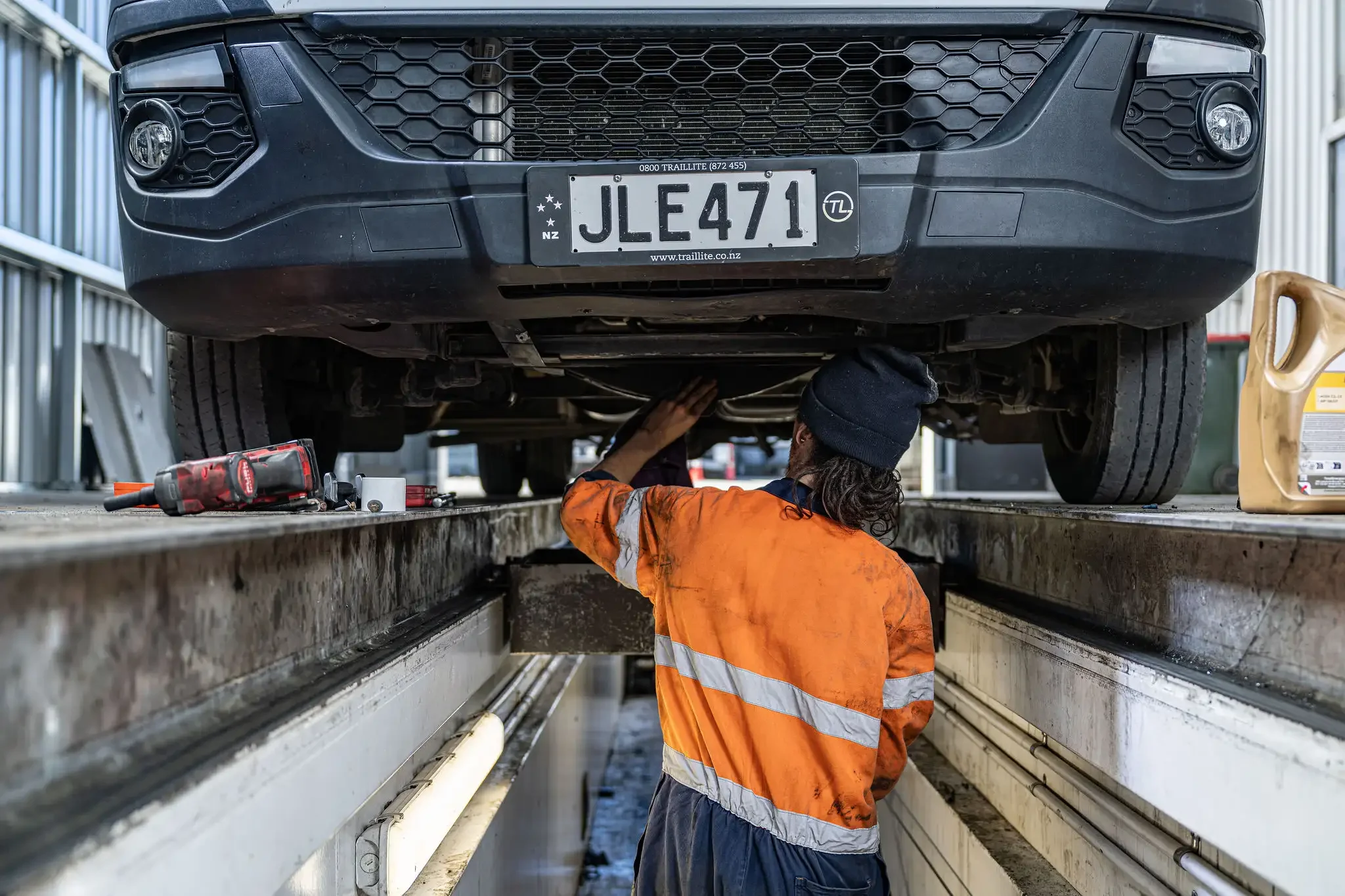 A mechanic in an orange high-visibility jacket working underneath a large dark-colored vehicle, likely a truck or SUV, that is elevated on a lift in an auto repair shop.