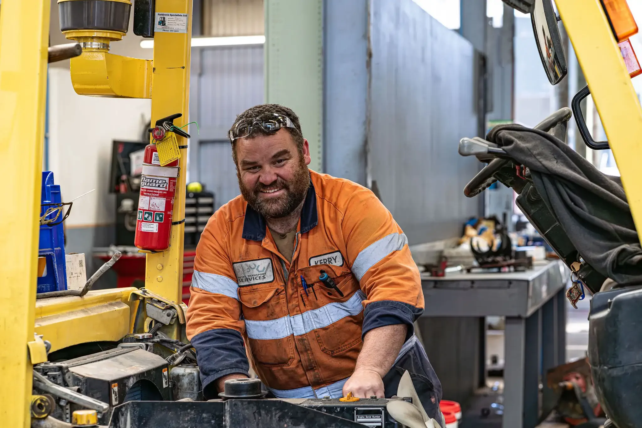 A man in a work uniform with orange and blue colors and a name badge kneels next to a vehicle engine in an automotive workshop. He is smiling and wearing safety glasses, surrounded by tools and equipment.