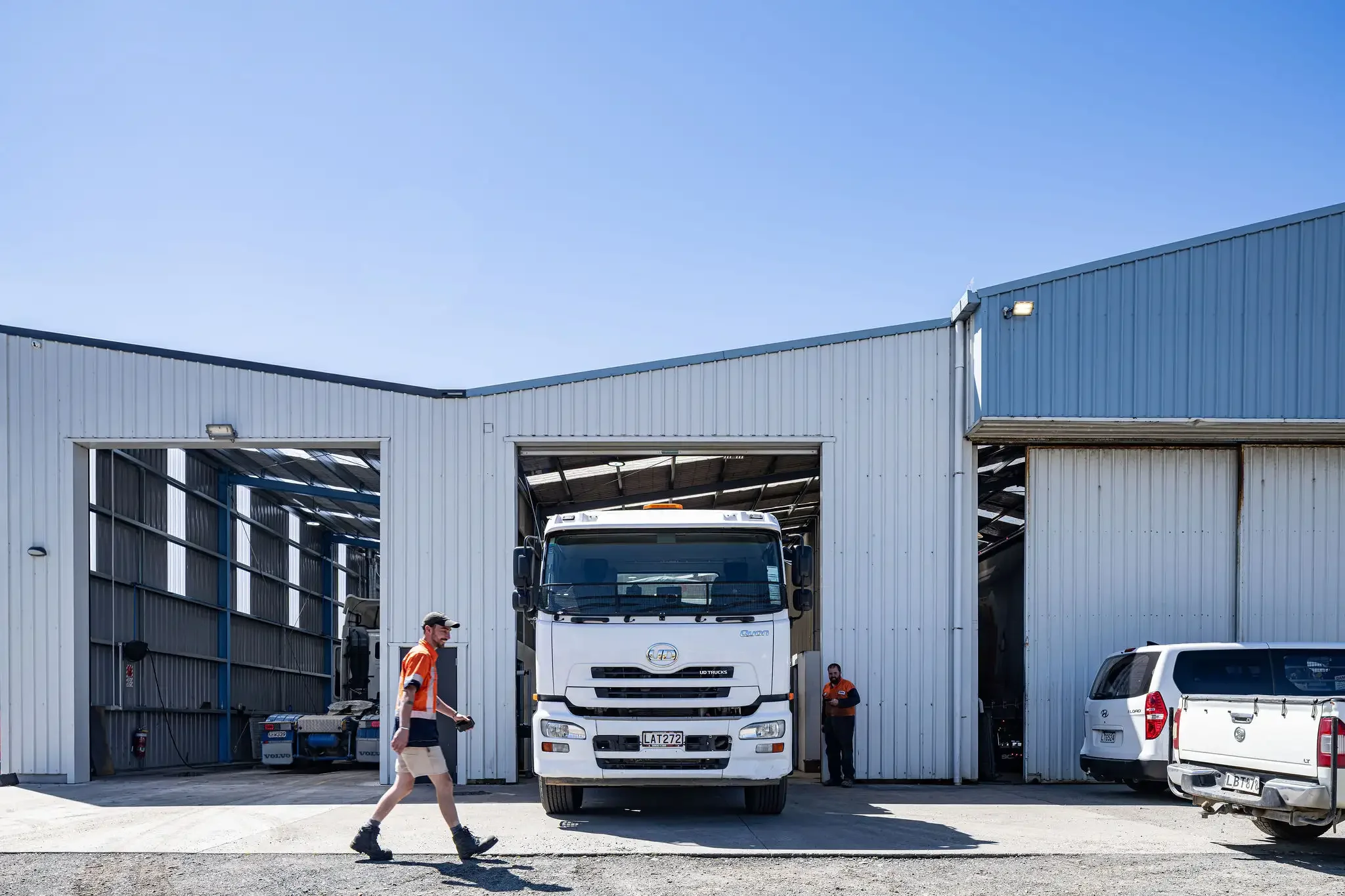 A white truck partially inside a large industrial building with two open garage doors, two workers in orange safety vests walking nearby, and a gray vehicle parked to the right in bright daylight.