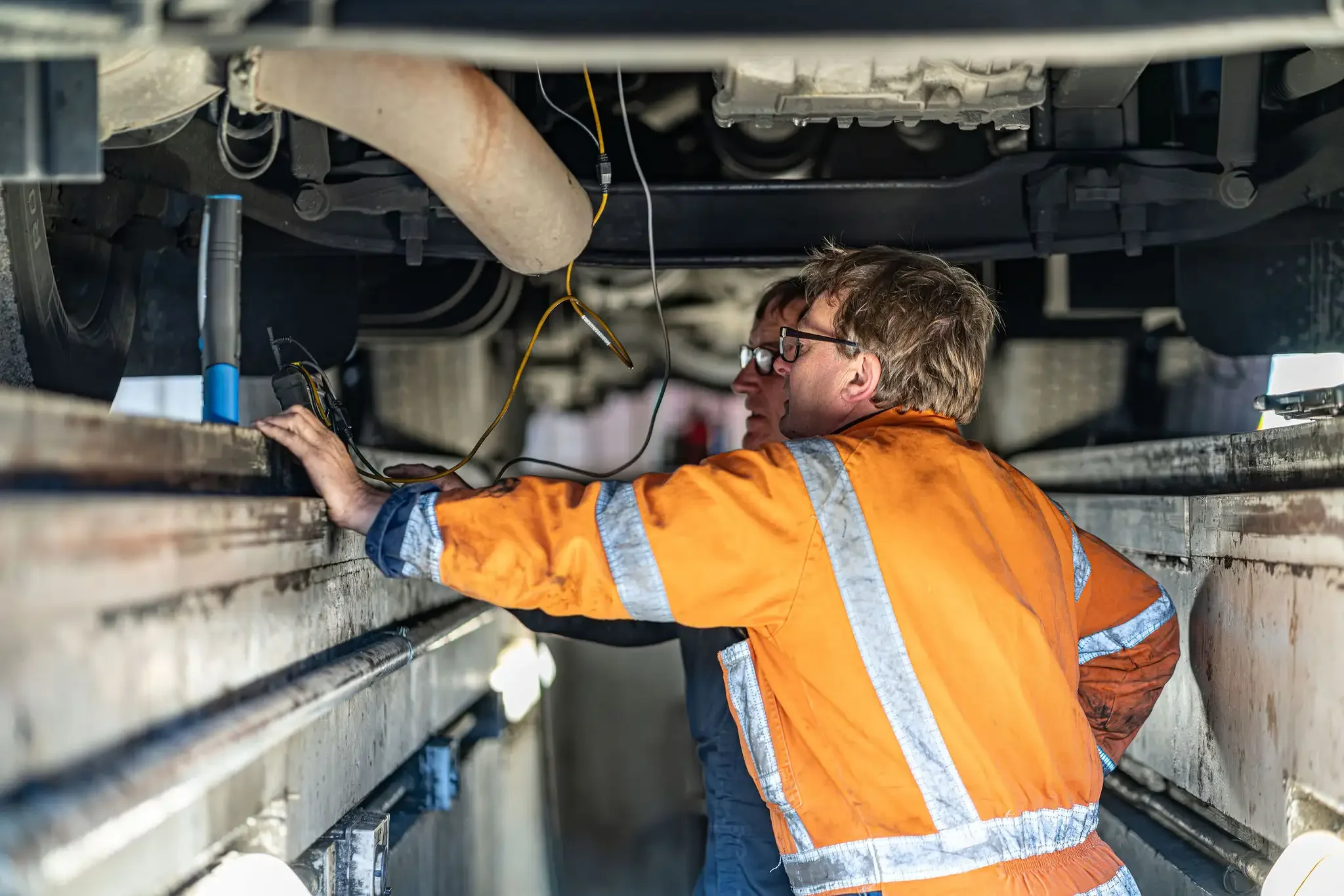 Construction workers in orange safety vests working underneath a large vehicle or machinery, inspecting or repairing it inside a metal framework.