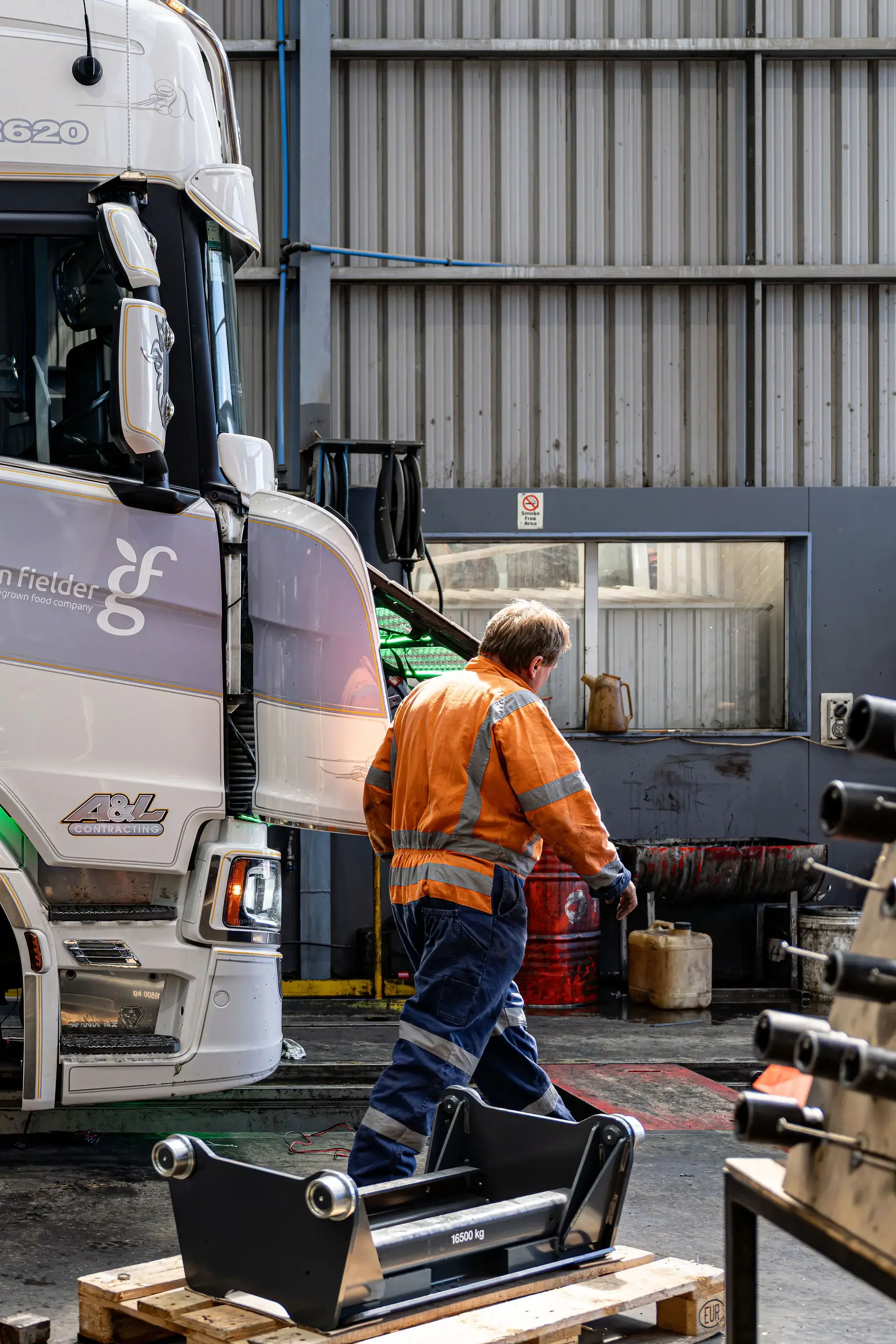 A worker in an orange safety jacket stands near a truck in an industrial or warehouse setting. The truck has branding that reads "field frog" and has a sticker indicating '48g Contracting.' The background features a metal wall with pipes, a window, and a "No Smoking" sign. The worker appears to be inspecting or working on equipment, with tools and machinery scattered around.