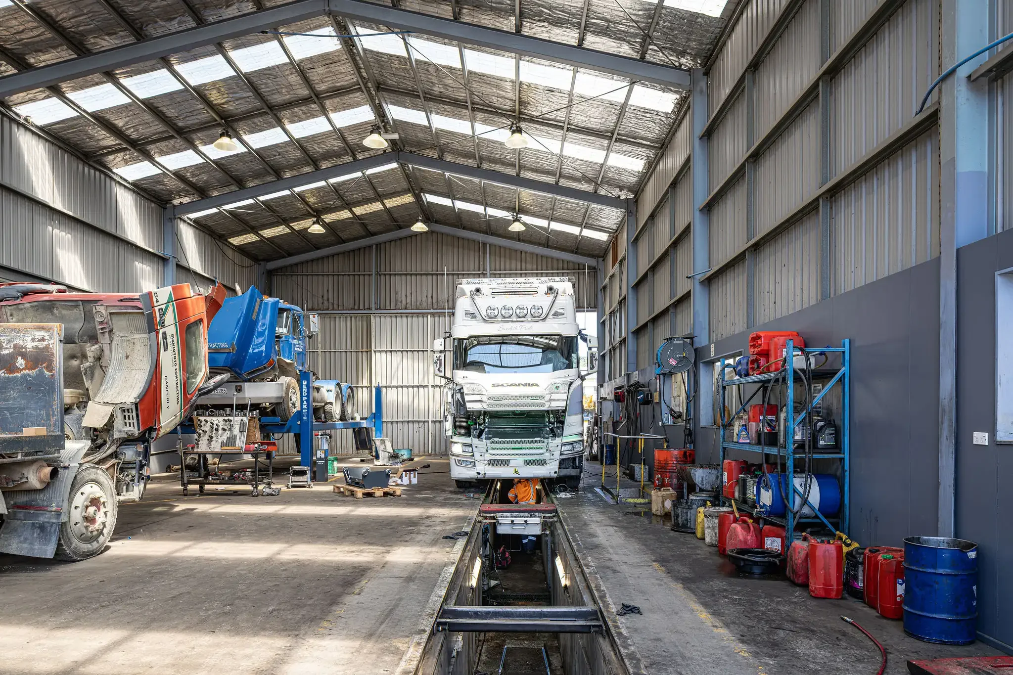 The interior of an auto repair shop with trucks and car parts, including a partially disassembled truck, a white truck on a lift, and various tools and equipment.