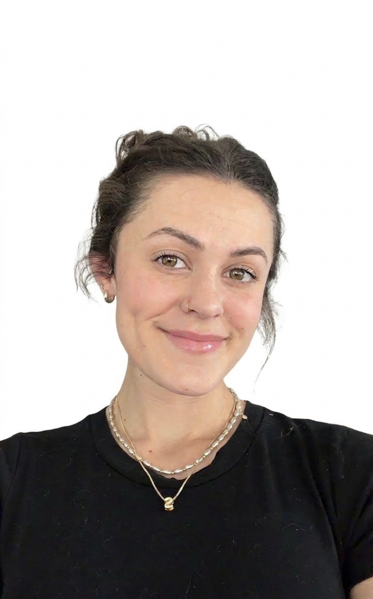 A woman with brown hair, light skin, and brown eyes smiling, wearing layered necklaces, hoop earrings, and a black shirt, standing against a plain white background.