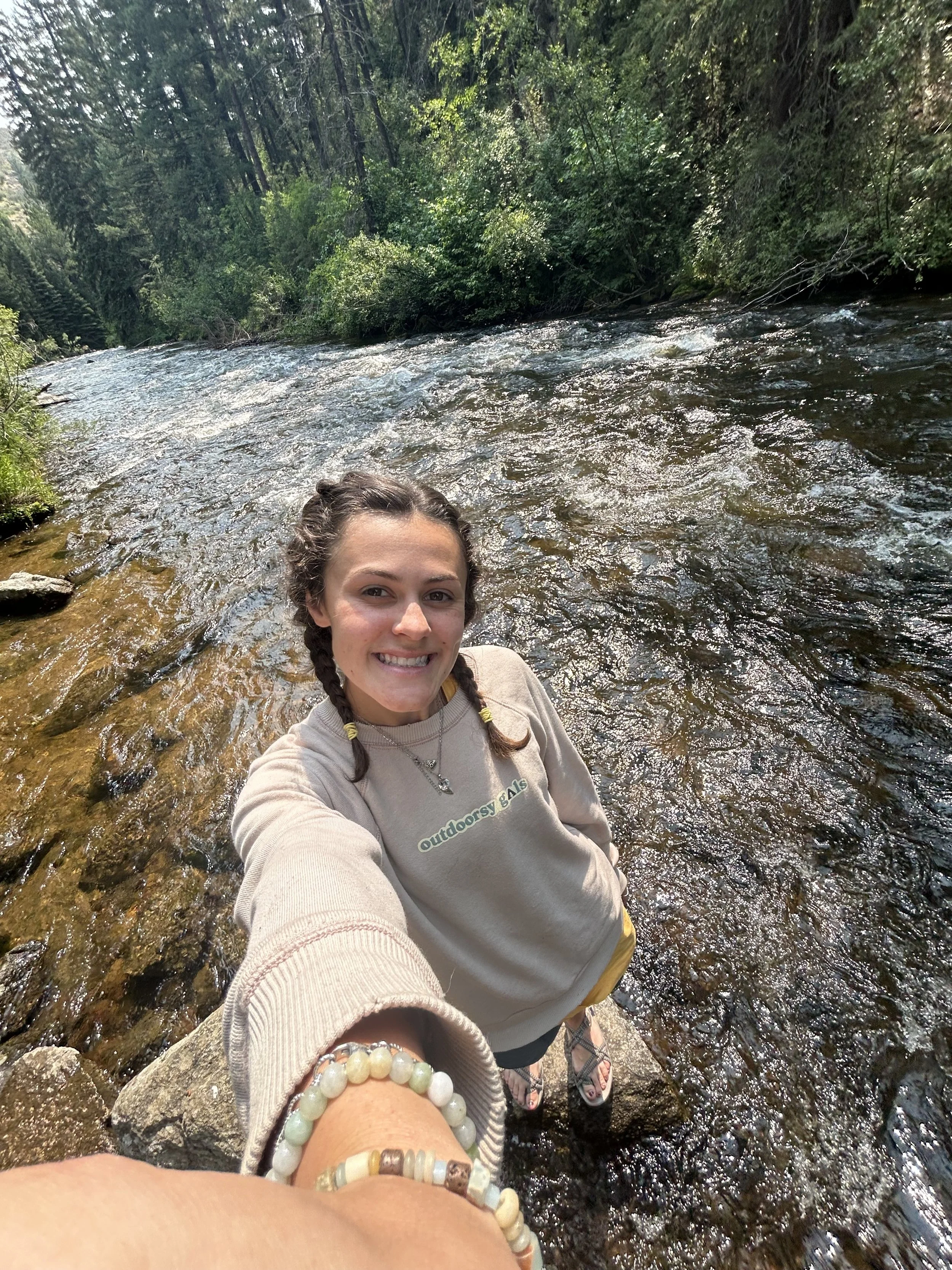 A smiling woman with braided hair wearing a beige sweatshirt that says 'outdoorsy gal' and jewelry, stands in a shallow, rocky river surrounded by trees and greenery. The photo appears to be taken from a high angle.