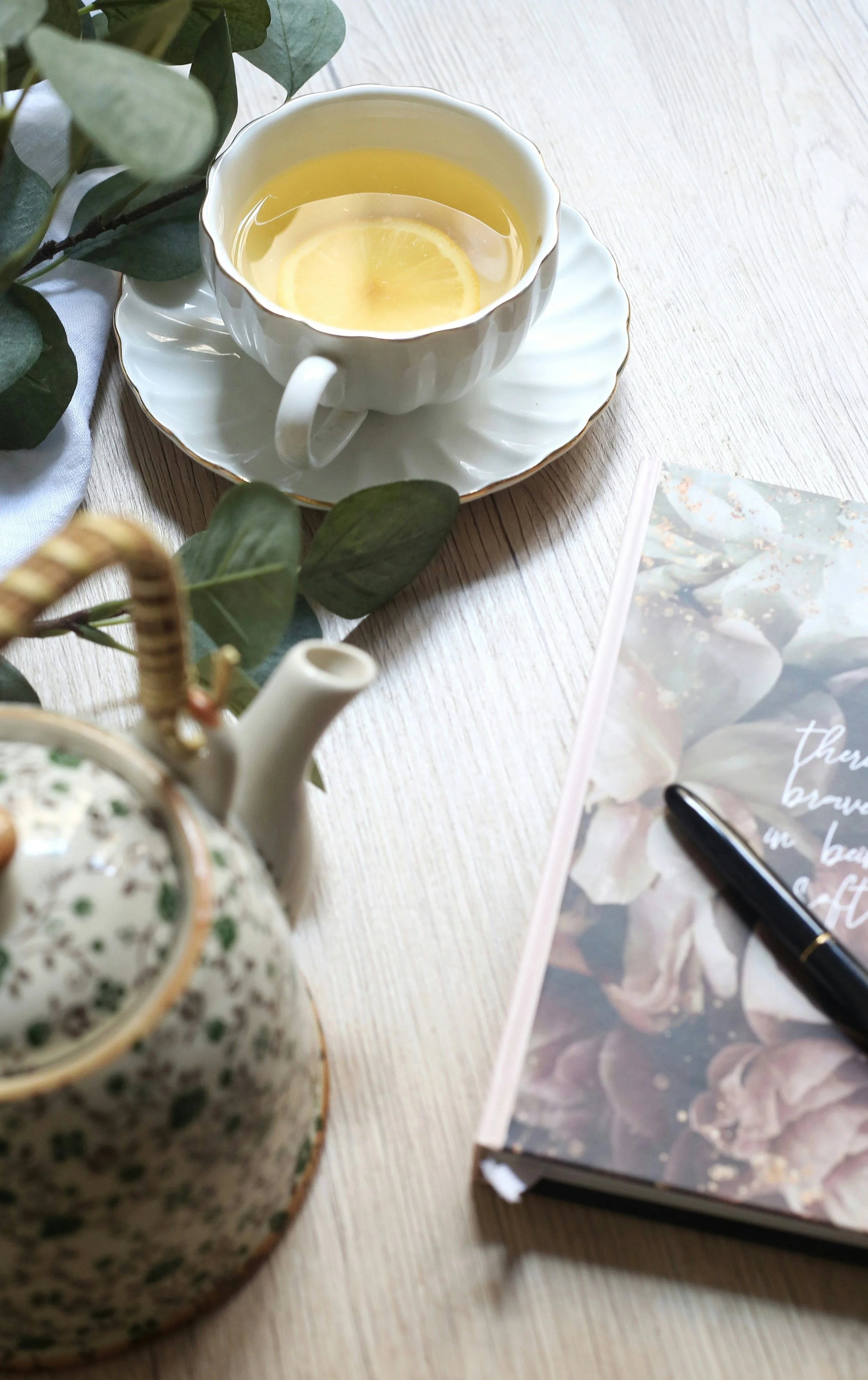 A teacup with lemon slices, a teapot with a floral pattern, a spiral-bound notebook with a floral cover, and a black pen on a light wooden table, with some green leaves.