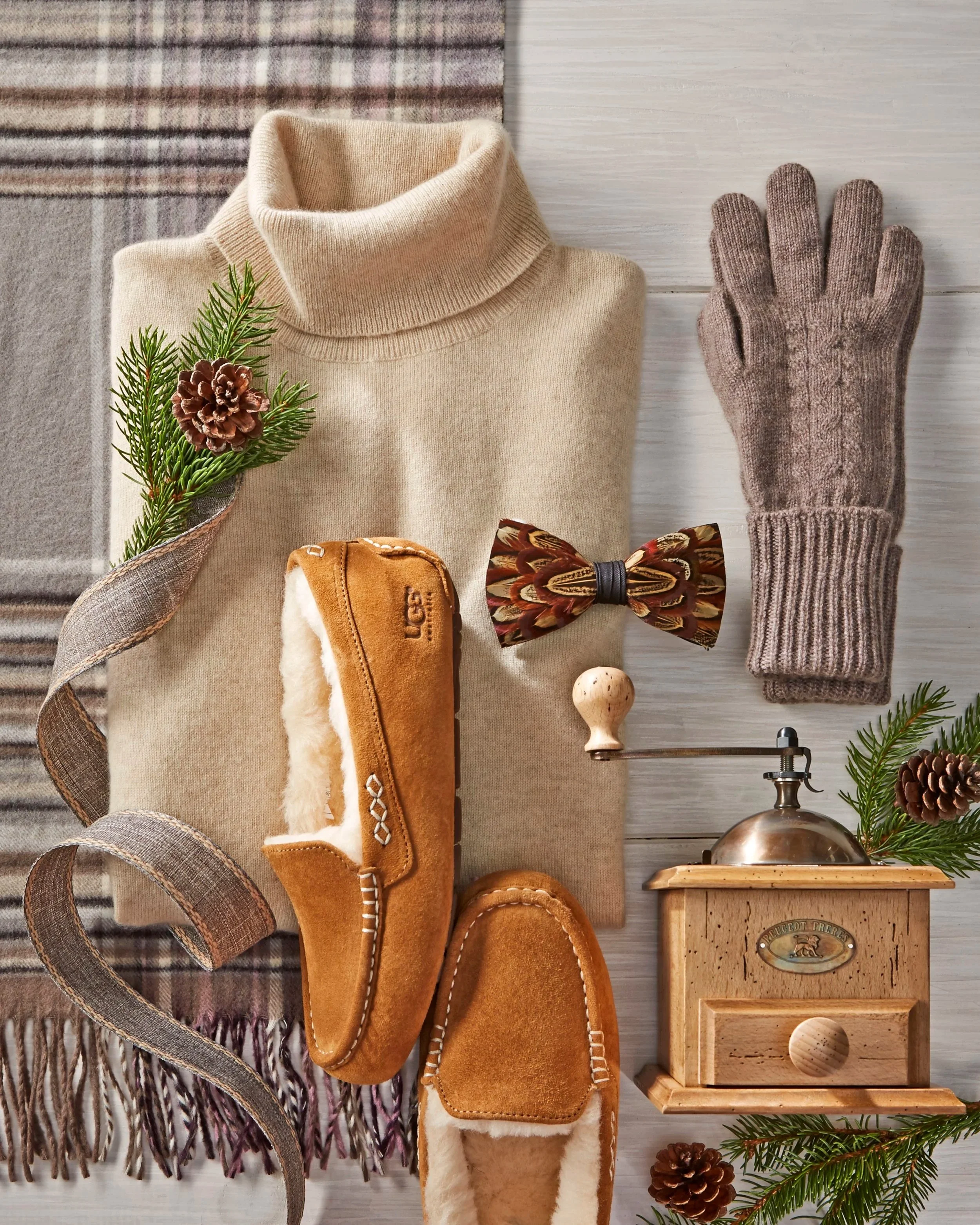 Flat lay of winter accessories and decorations including a beige turtleneck sweater, brown gloves, plaid scarf, brown slippers, a wooden caddy with a metal handle, pinecones, sprigs of pine, and a bow tie on a light wooden surface.