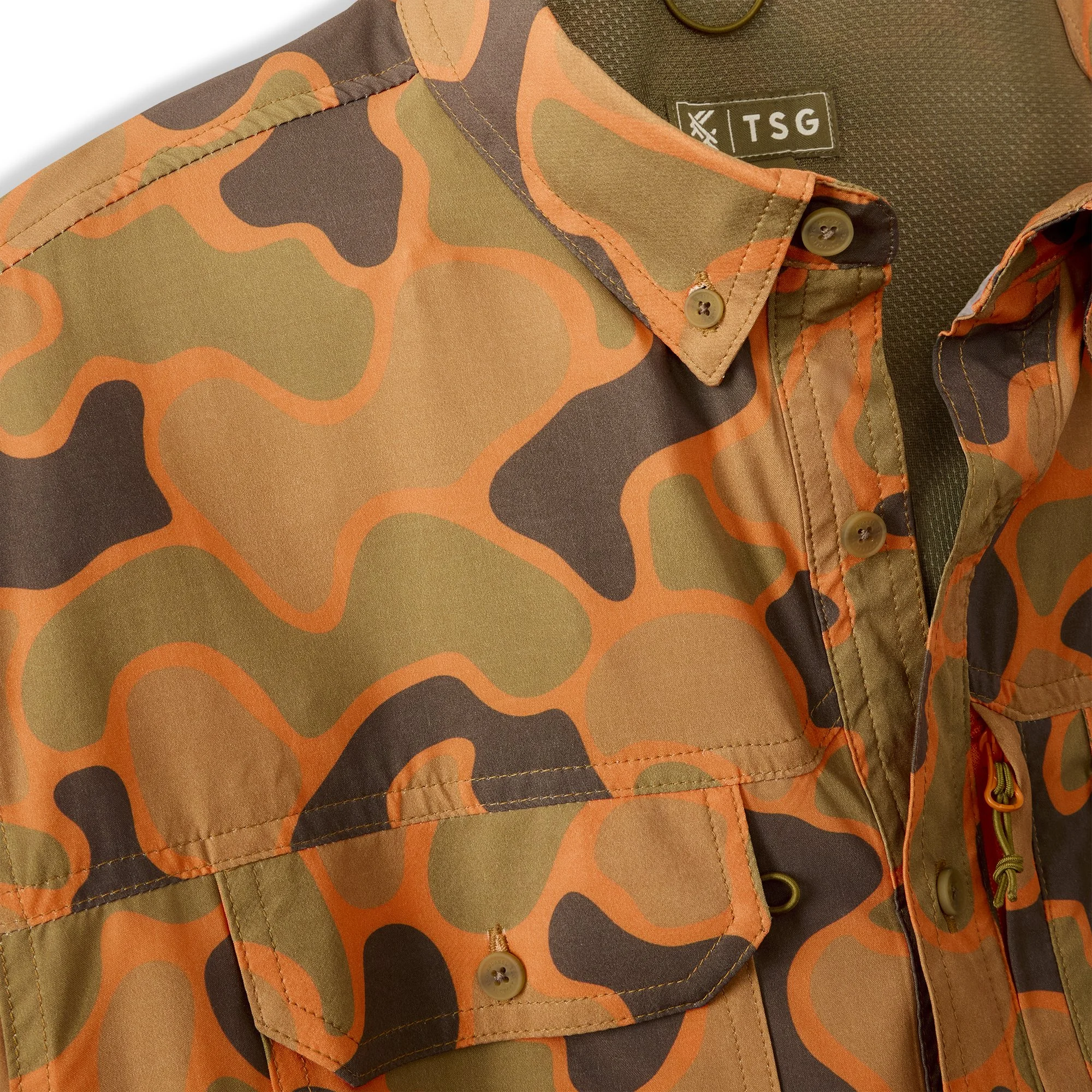 Close-up of a camouflage jacket with orange, green, brown, and black patterns, featuring buttons, stitching, and a label with the letters 'TSG'.