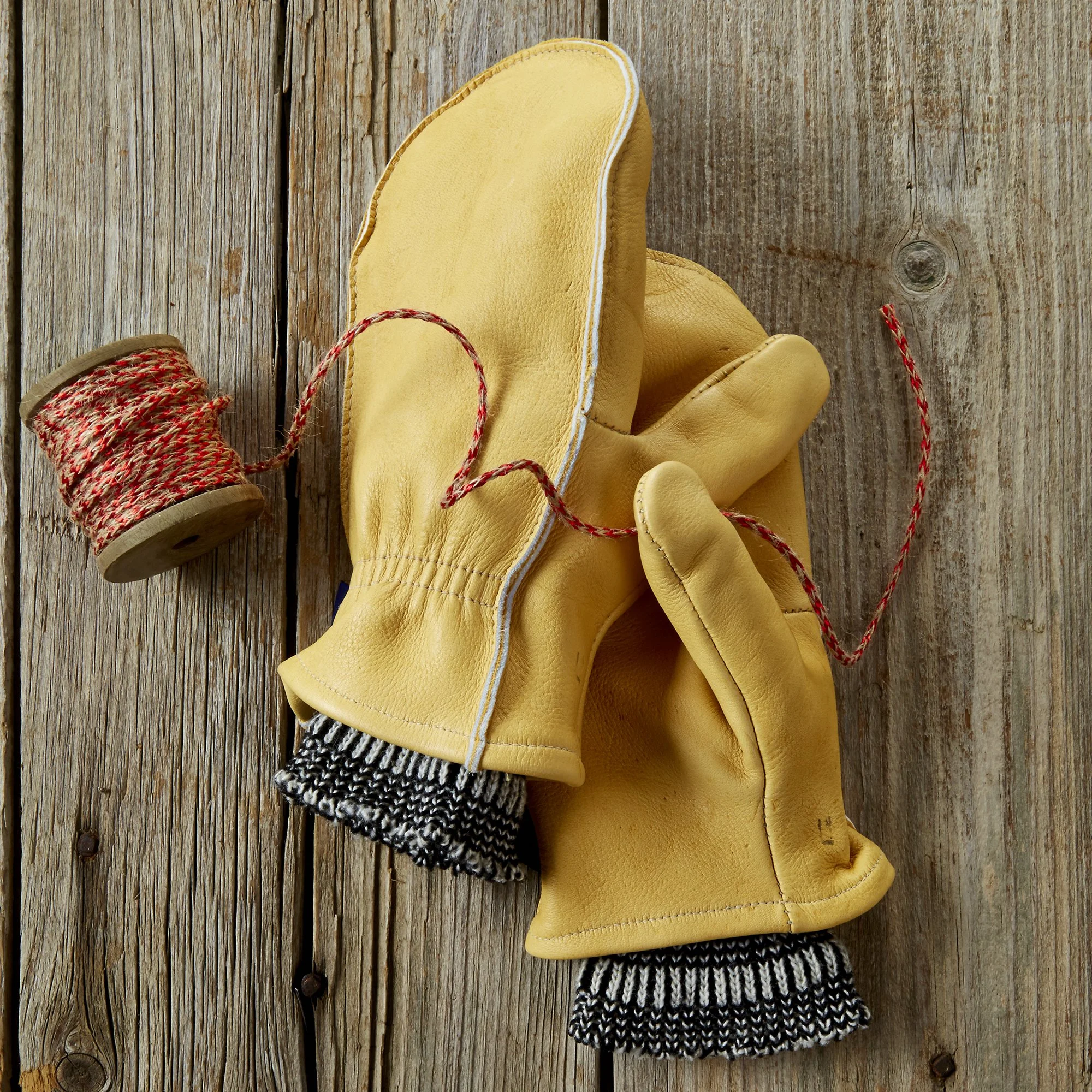 Yellow work glove with a leather exterior, a knit cuff, and red and white drawstrings on a wooden surface.