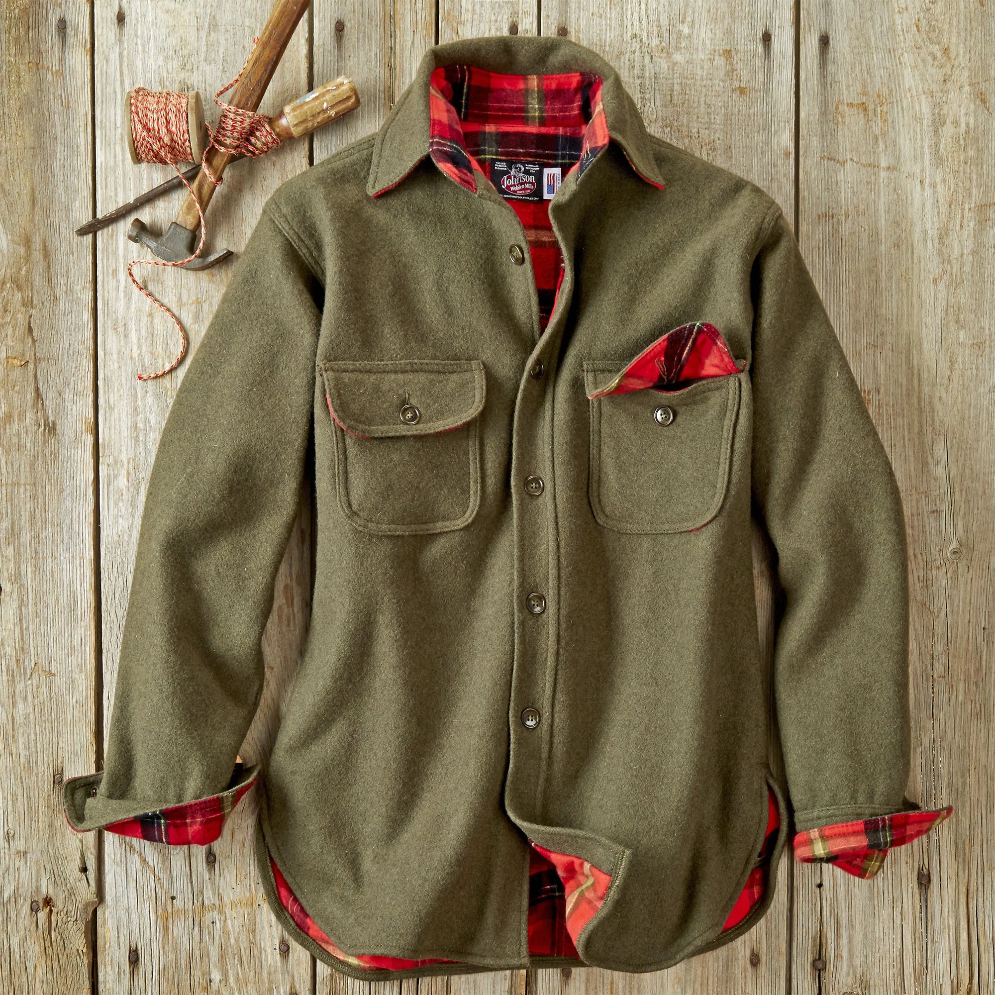 A green outdoor jacket with red plaid lining and pockets, hanging on a wooden wall with a hammer and red and white string nearby.