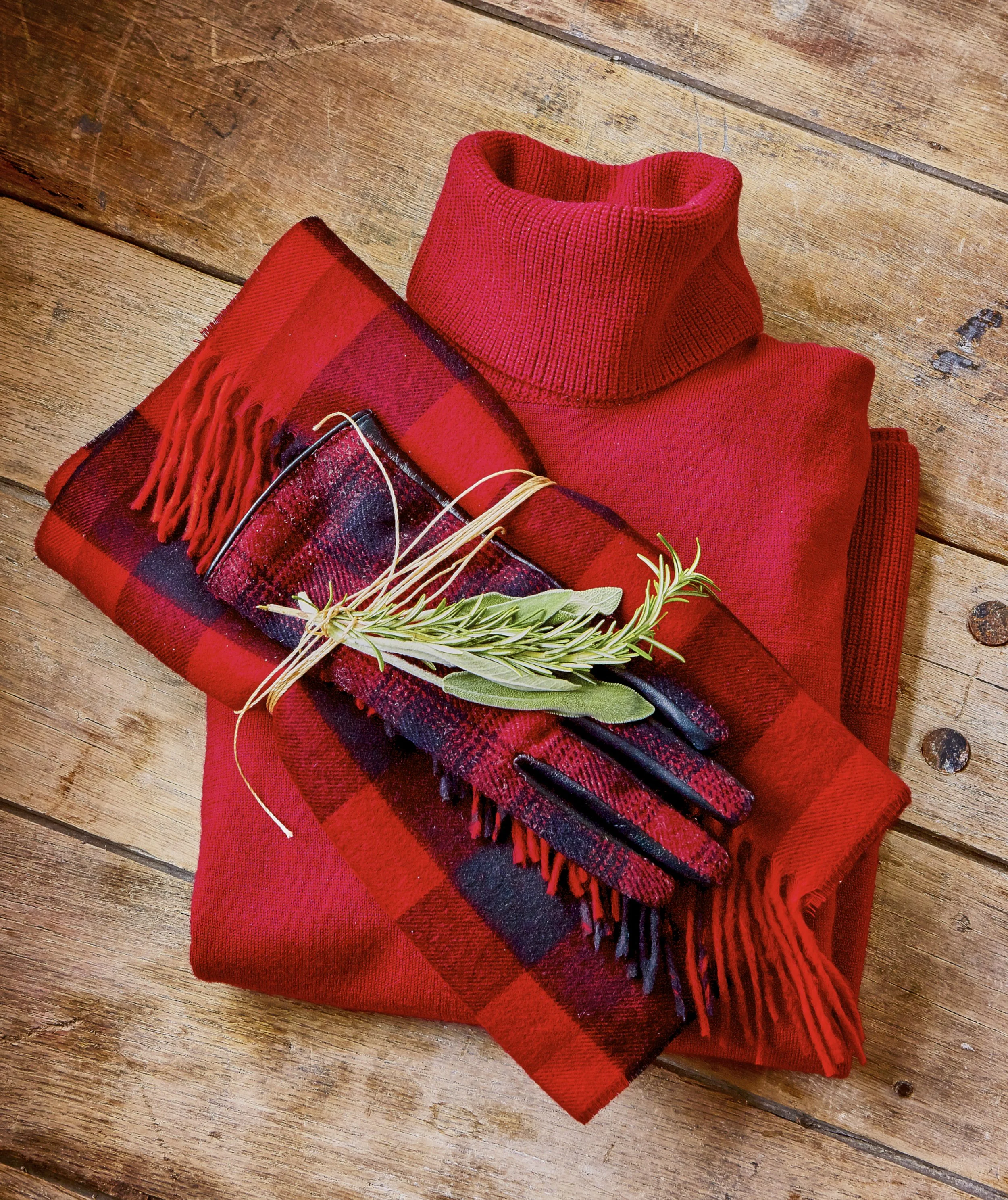 A red turtleneck sweater and a red plaid scarf wrapped with a beige string and a sprig of green leaves, placed on a wooden floor.