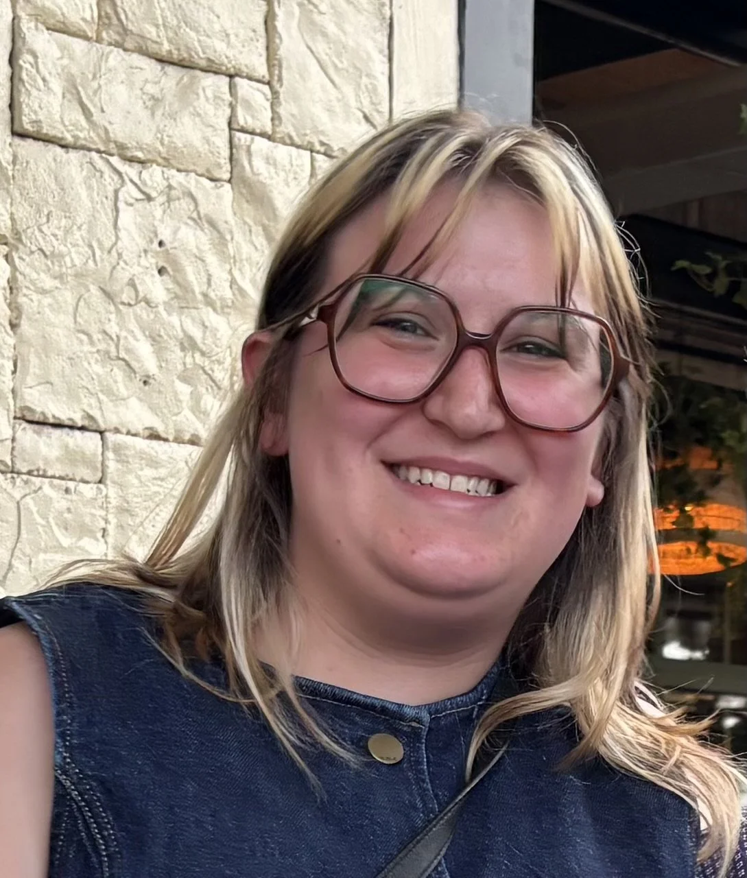 Smiling woman with glasses, wearing a sleeveless denim top, in front of a stone wall at dusk.