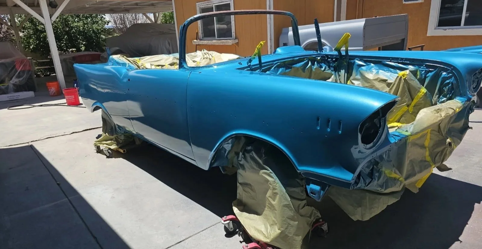 A vintage car in process of being painted, covered with blue paint. The car is partially masked with paper and tape for painting, and is mounted on wheel dollies in a garage or outdoor workspace.