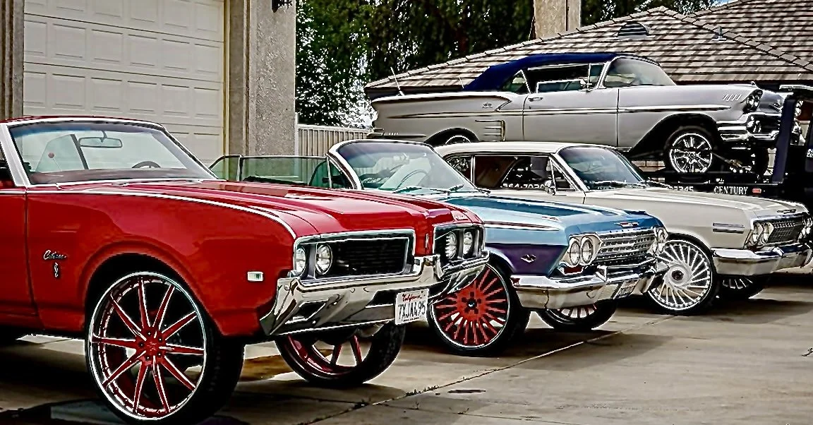 Four classic cars parked in a driveway, with a garage and a house in the background.