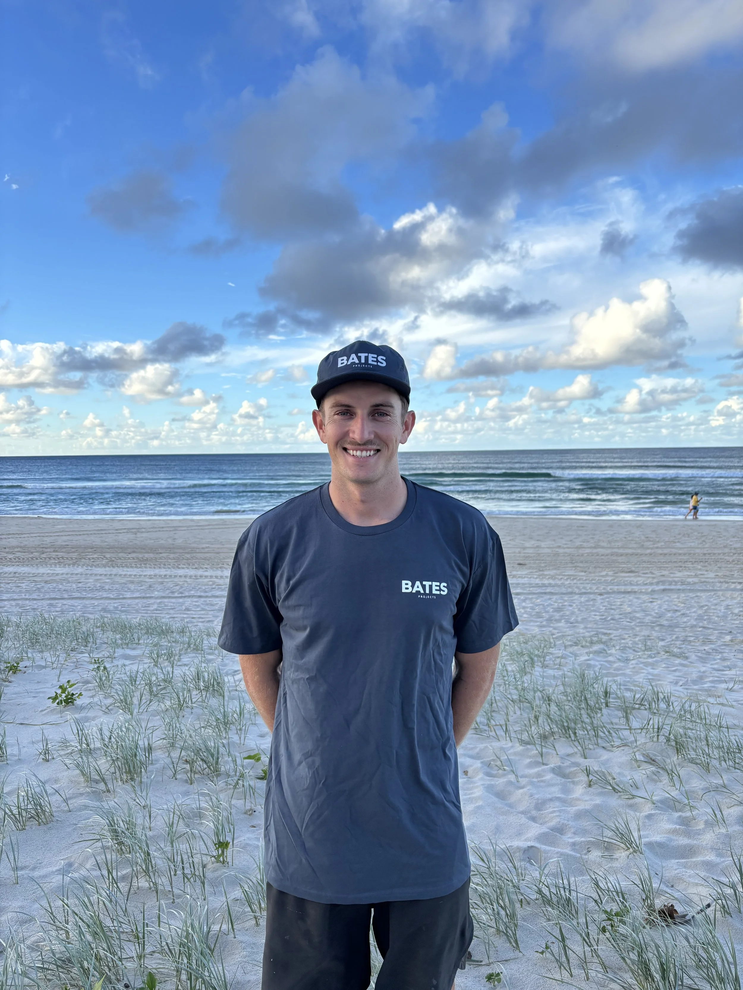 A young man in a dark blue BATES t-shirt and matching cap smiling at the camera on a sandy beach with grass, ocean, and a partly cloudy sky in the background.