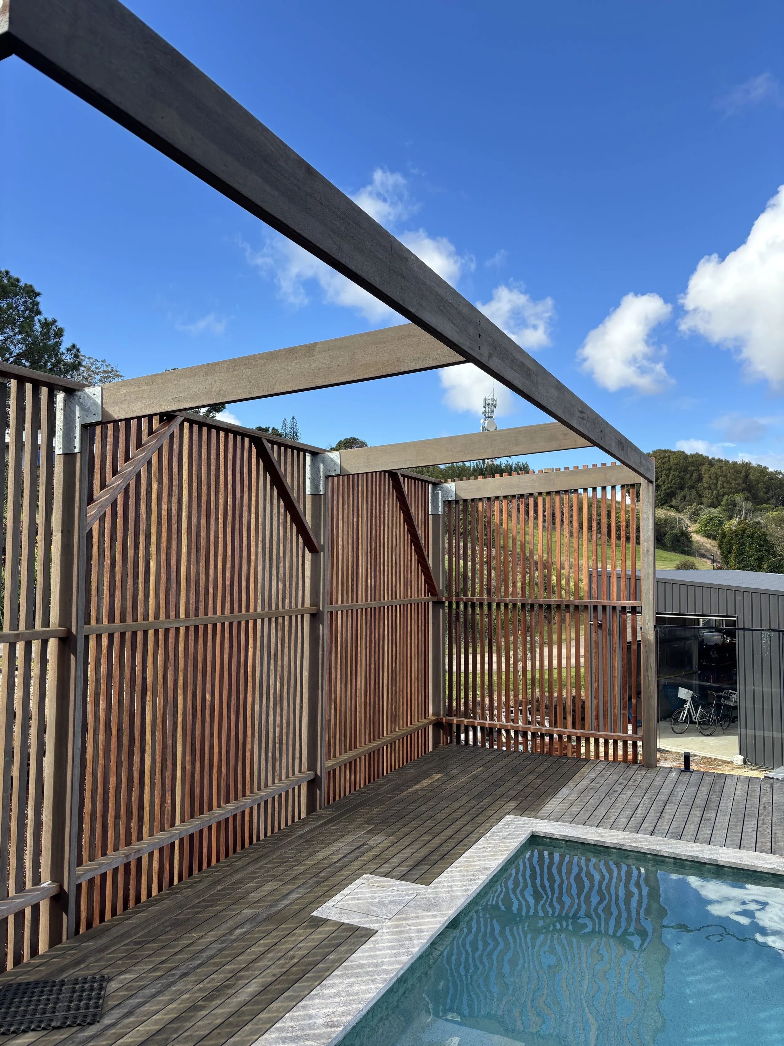 Partially constructed wooden fence and frame around a small swimming pool on a sunny day with blue sky and clouds.