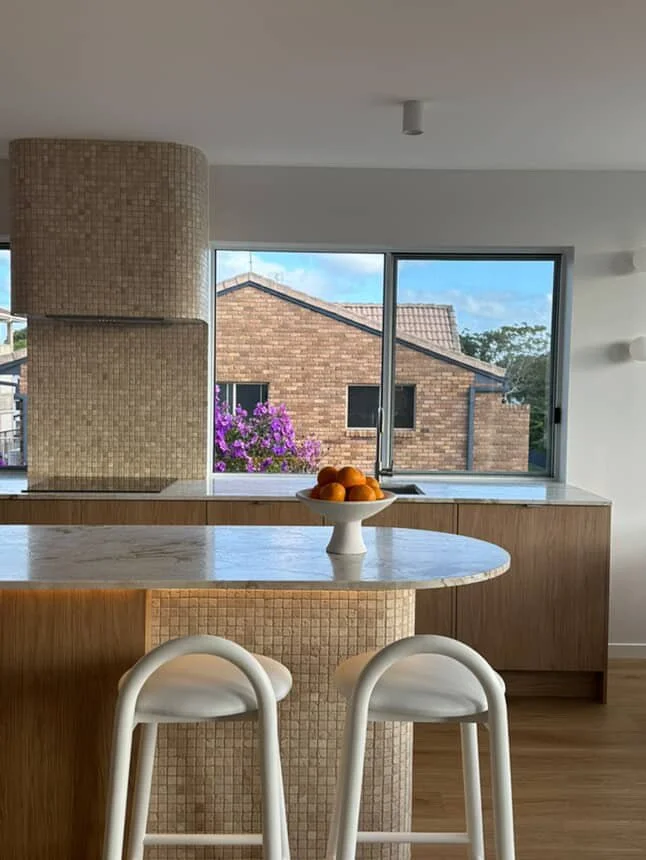 Interior view of a kitchen with a marble countertop island, two white chairs, a bowl of oranges, a large window showing a neighboring house with a brick exterior, and a purple flowering tree outside.