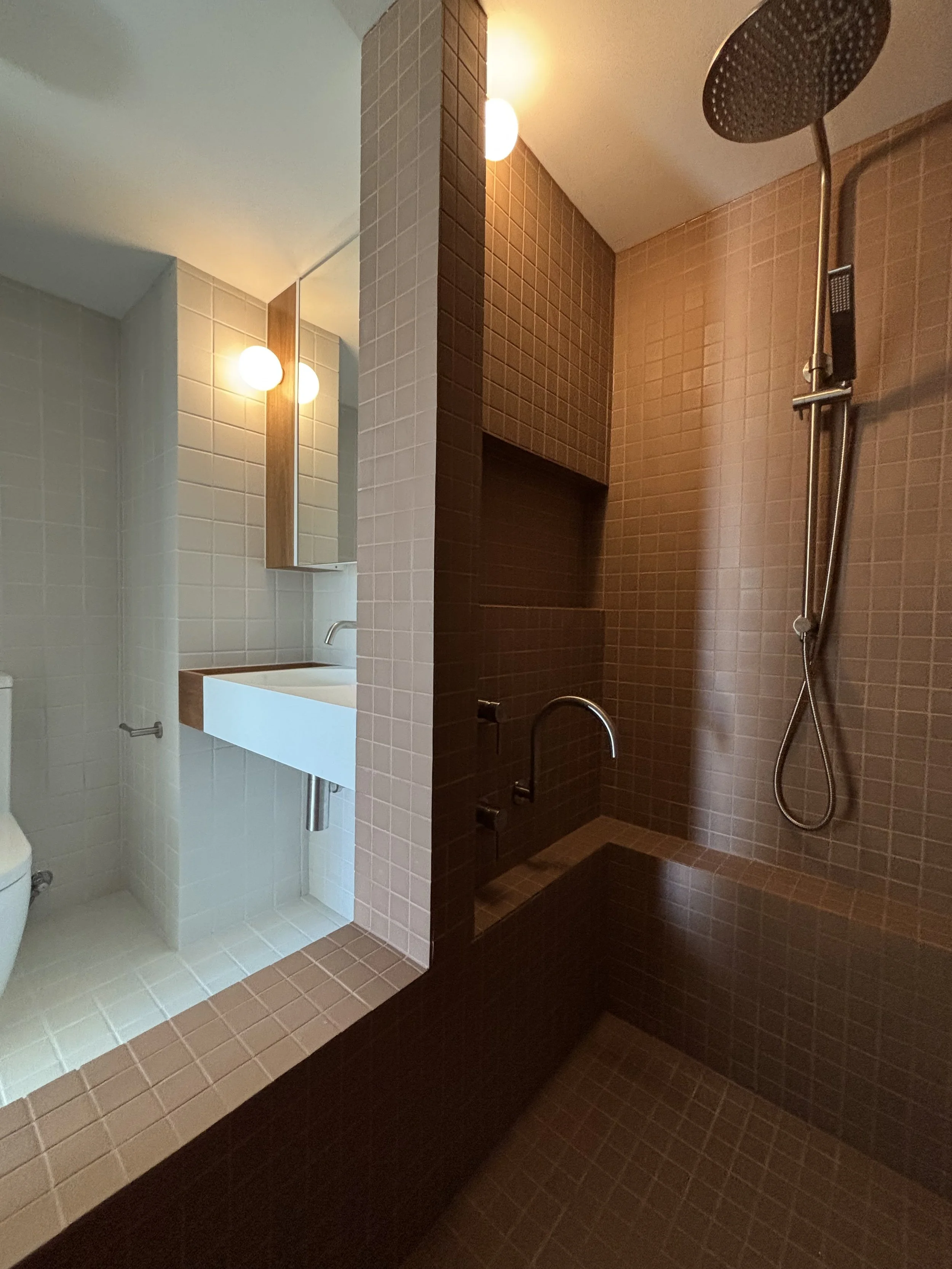 A bathroom with a separate shower area with dark brown tiles and a rain shower head, and a white sink with a mirror and round light fixtures, divided by a partial wall.