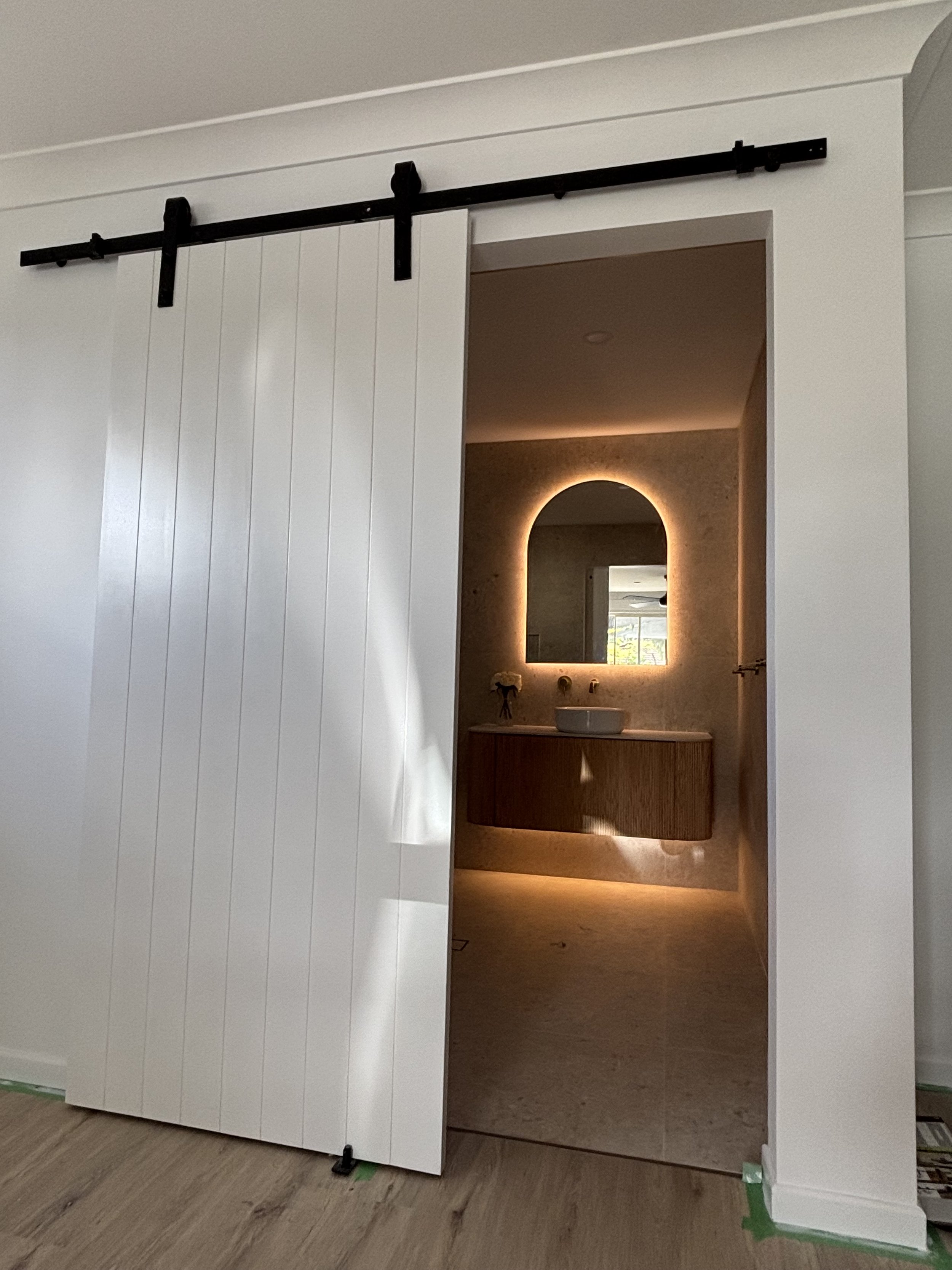 A sliding barn door partially open revealing a modern bathroom with a backlit mirror, a vessel sink, and a wooden vanity.